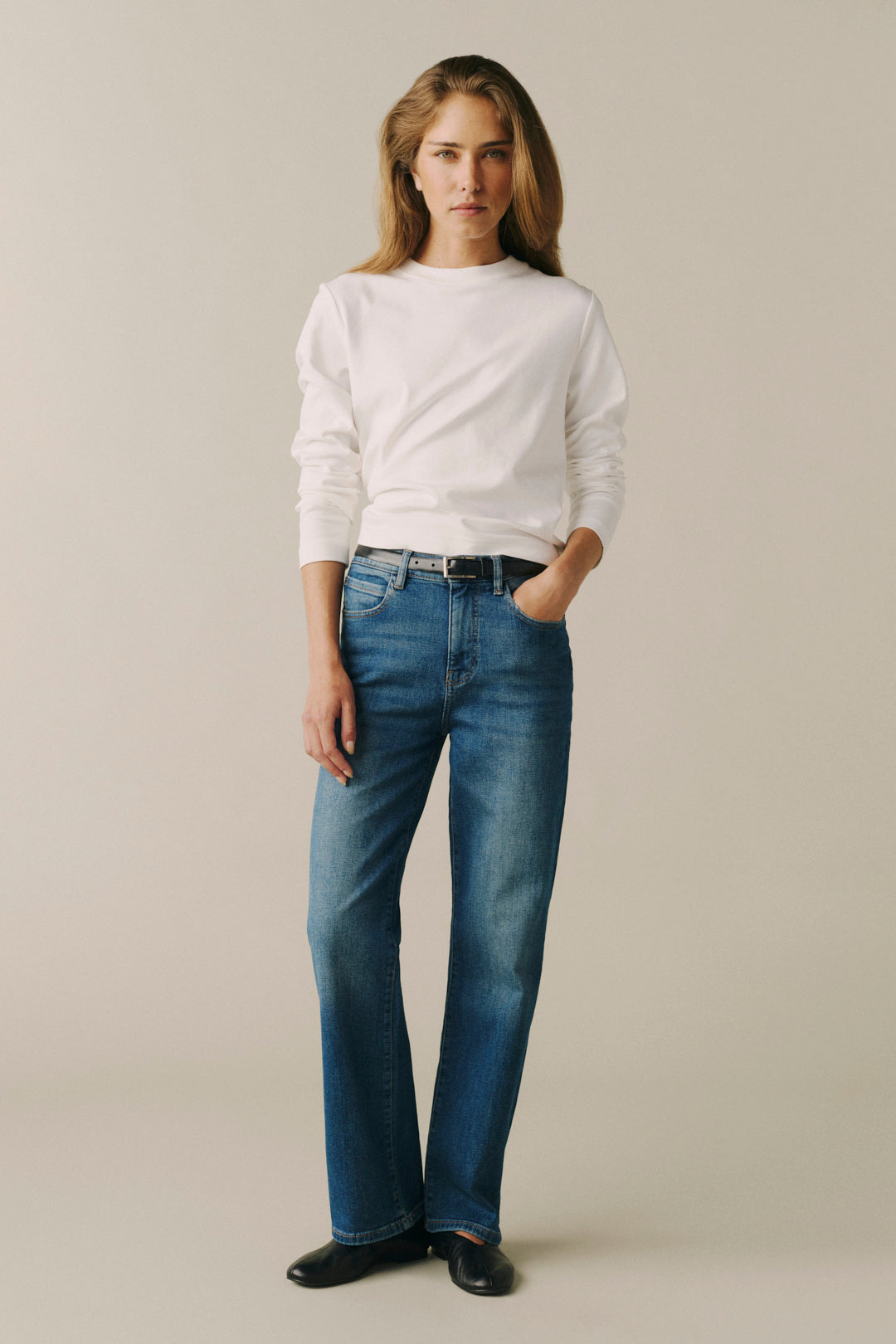 A woman modeling a white long-sleeve top and blue straight-leg jeans against a plain beige studio background.