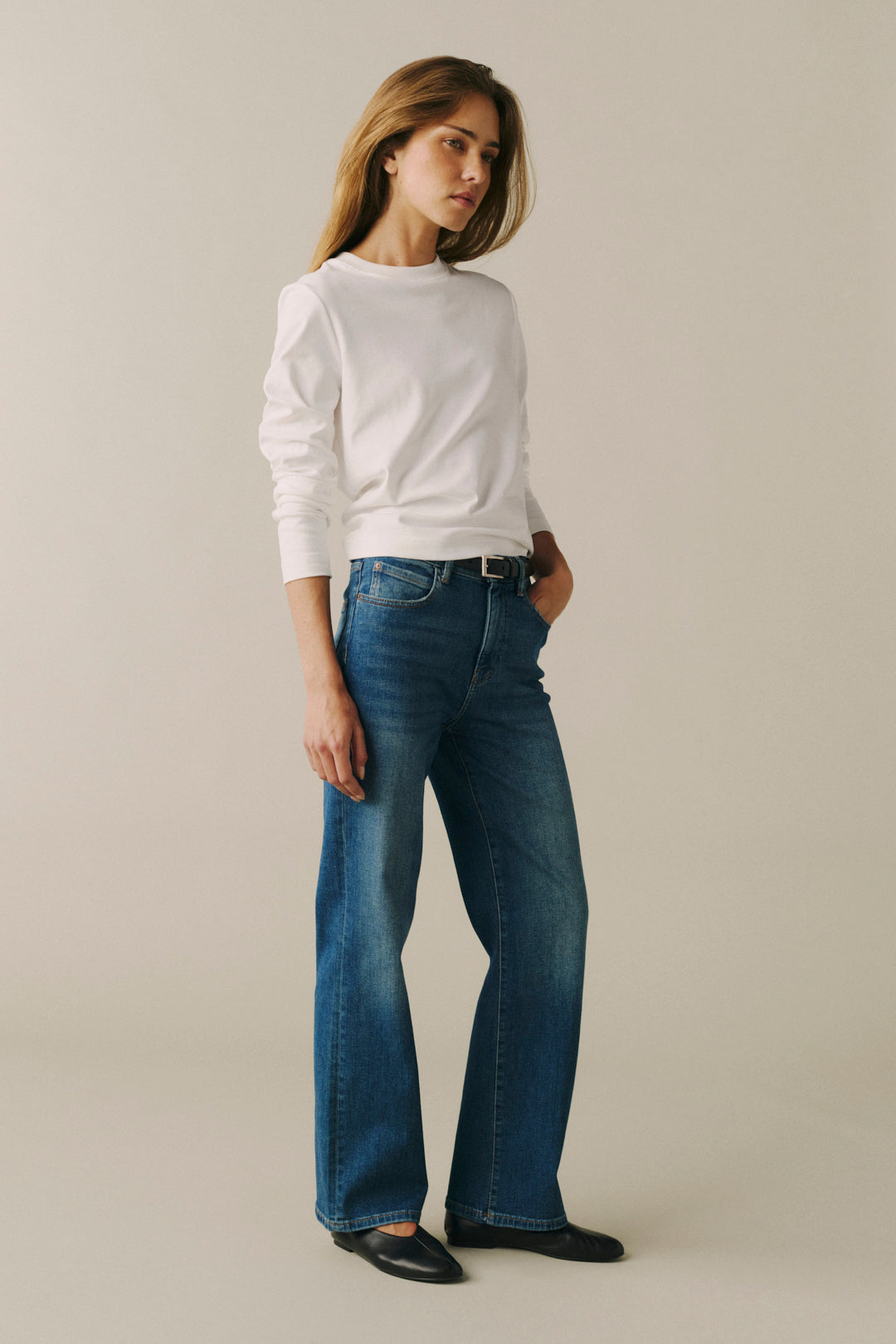 A woman modeling a white long-sleeve crewneck top and blue wide-leg jeans against a plain beige studio background.