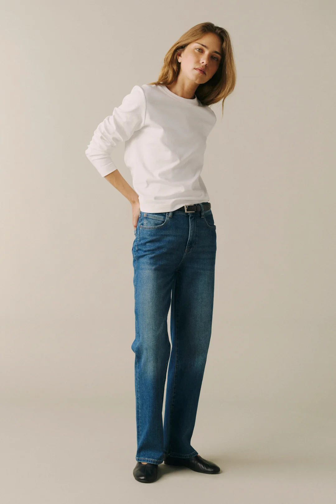 A woman modeling a white long-sleeve crewneck shirt and blue straight-leg jeans with black shoes, standing against a plain light beige studio background.