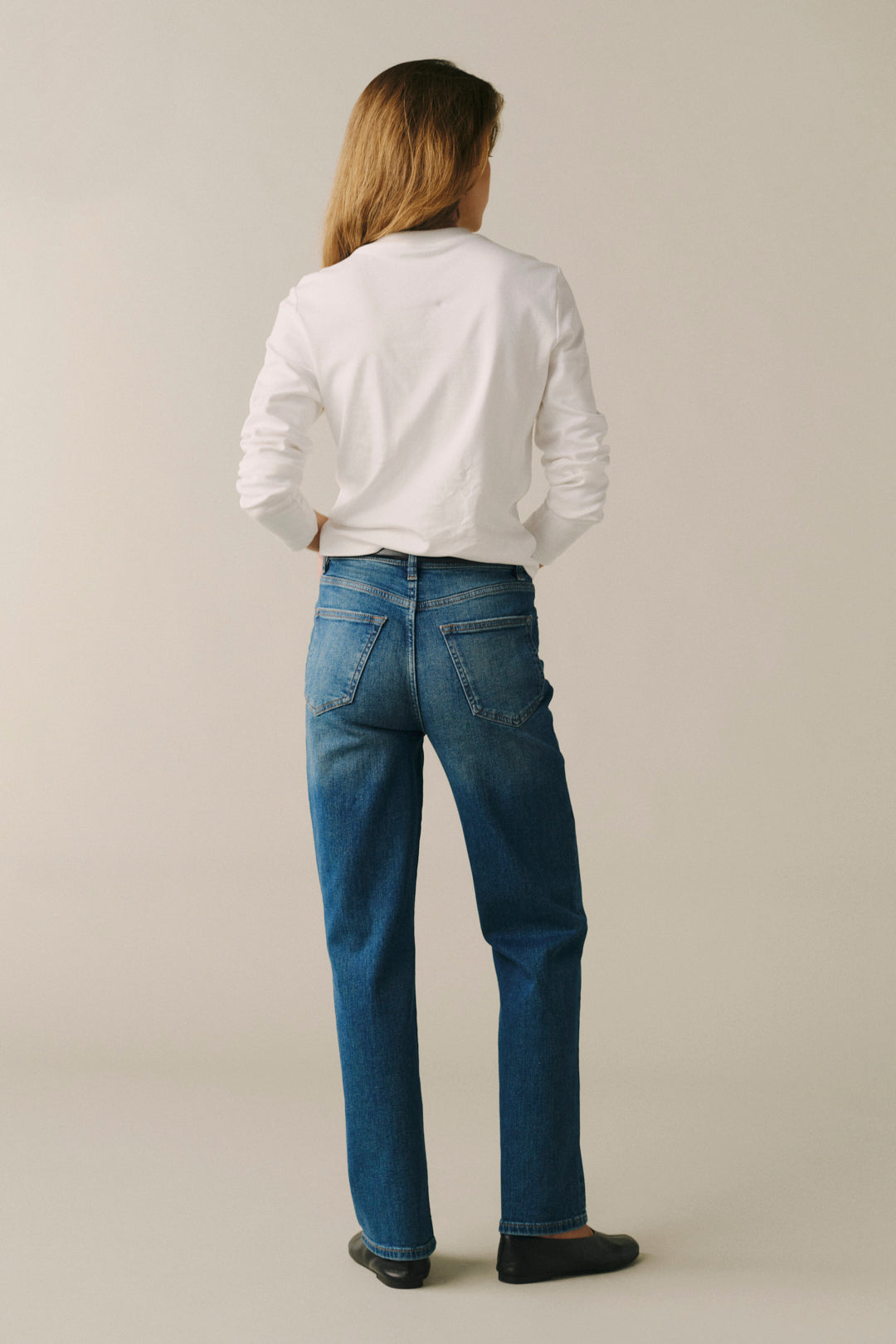 A woman is shown from behind wearing a white long-sleeve button-up shirt and blue straight-leg jeans against a plain light beige studio background.