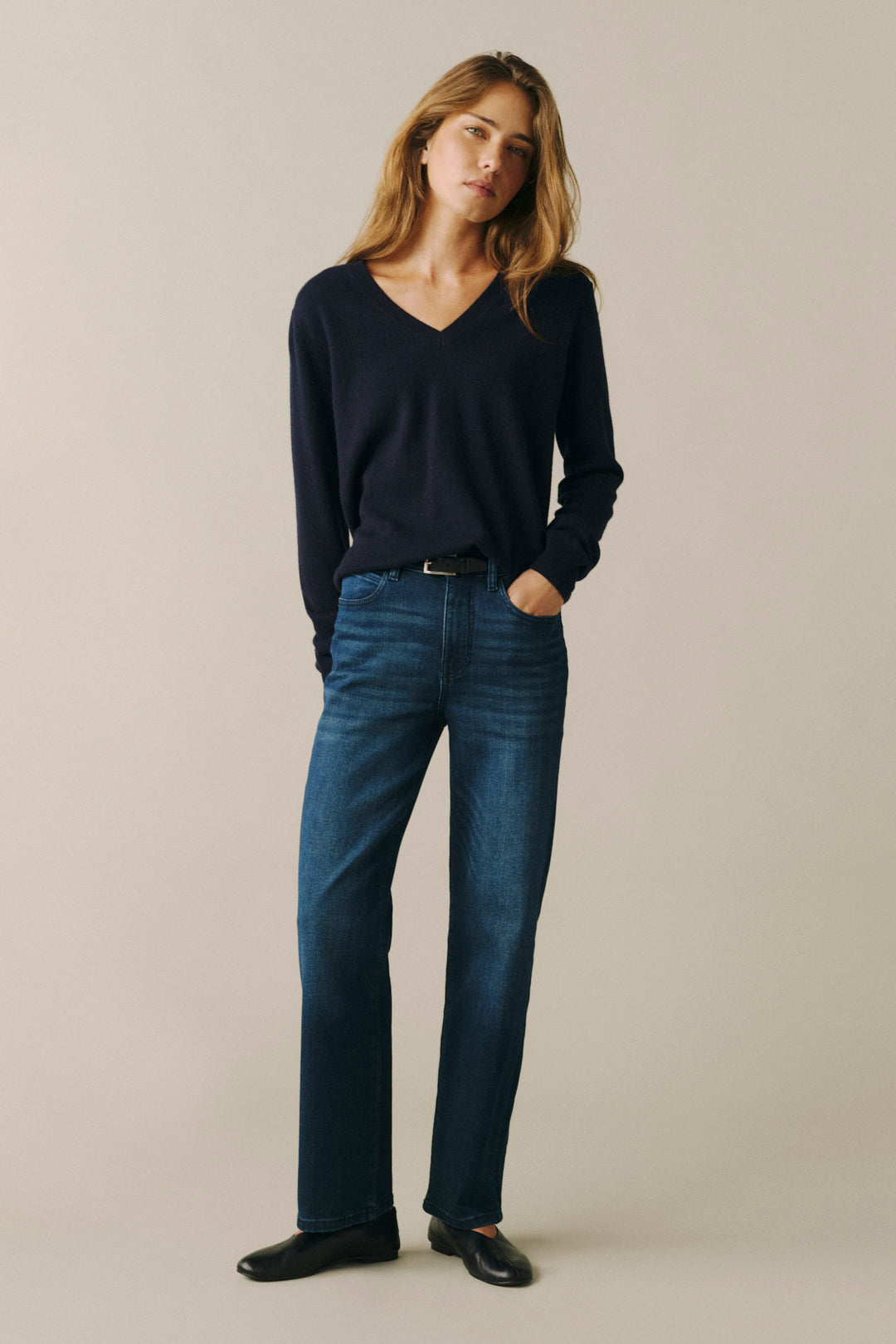 A woman modeling a dark navy blue long-sleeve V-neck sweater and dark blue straight-leg jeans, paired with black shoes, against a solid light beige studio background.