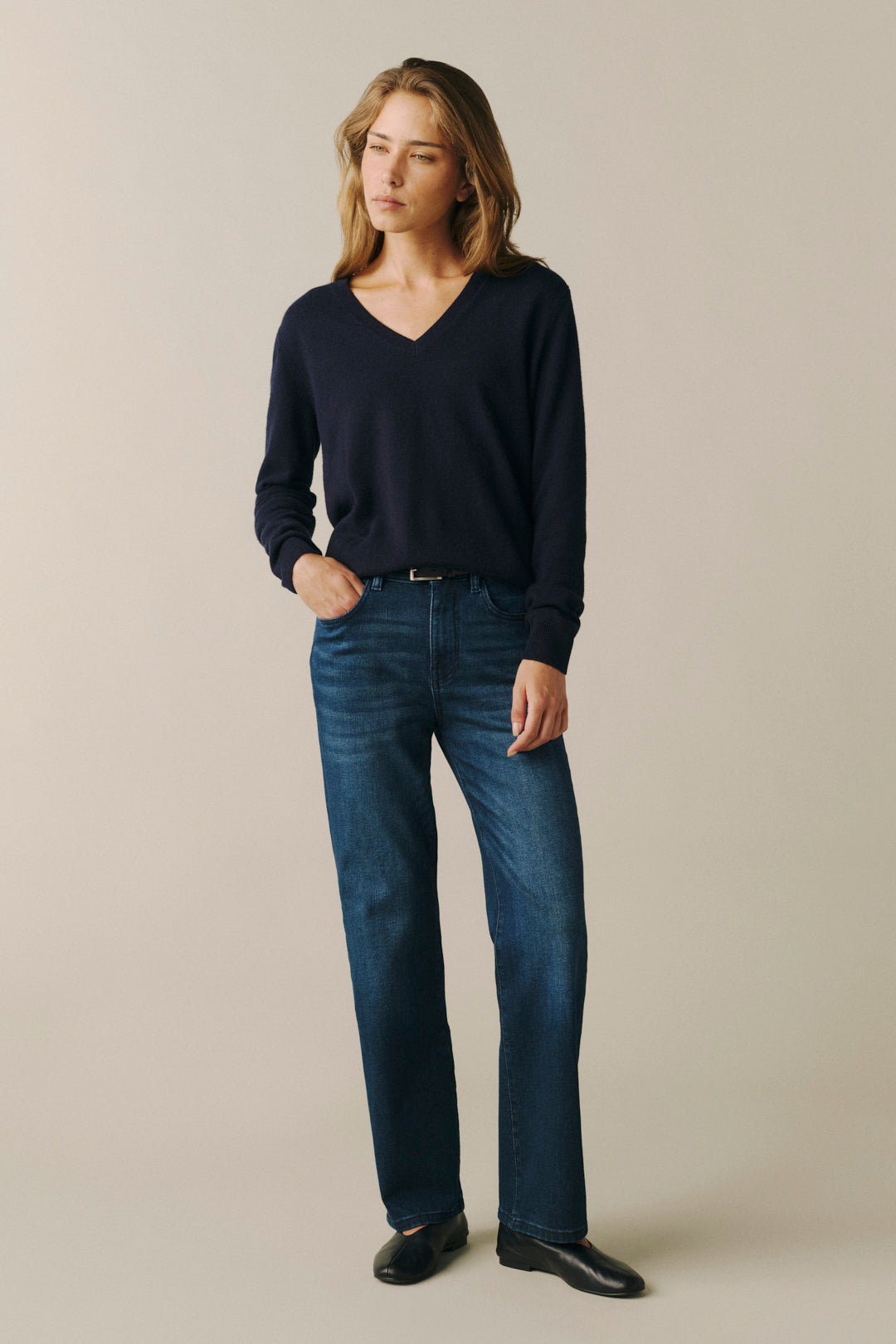 A woman modeling a dark navy blue V-neck knit sweater and dark blue straight-leg jeans, standing against a solid light beige studio background.