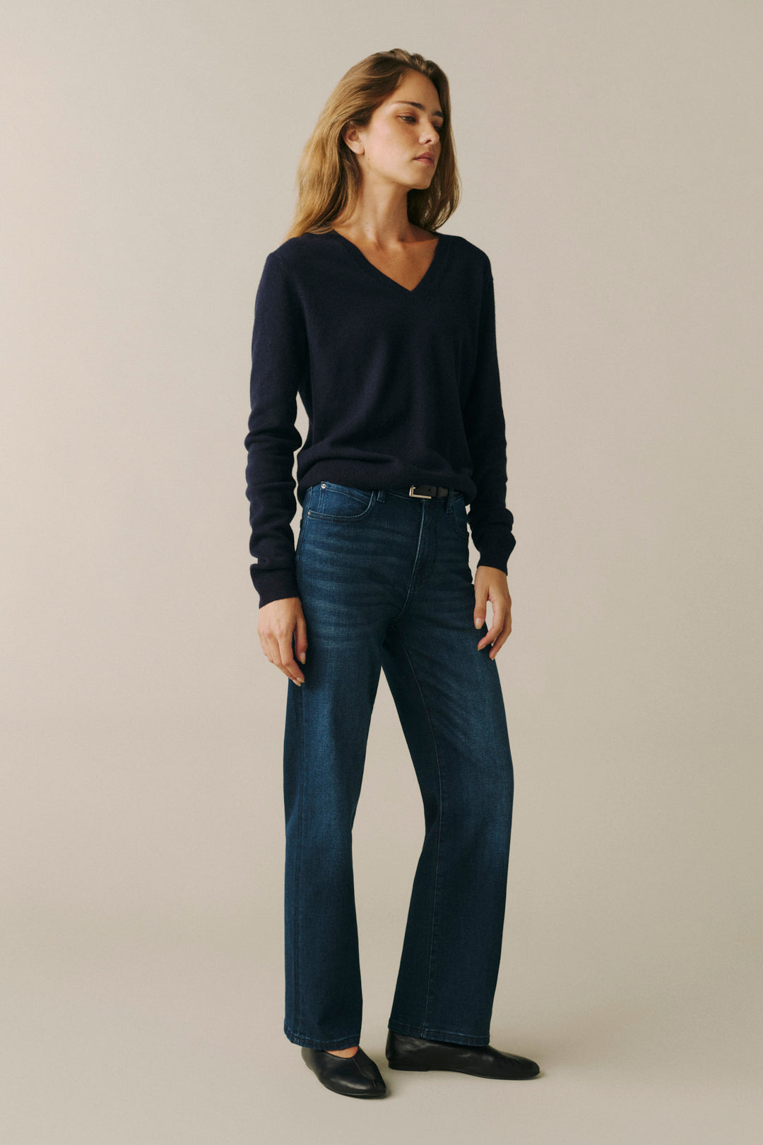 A woman modeling a dark navy long-sleeve V-neck sweater and dark blue straight-leg jeans against a plain beige studio background.