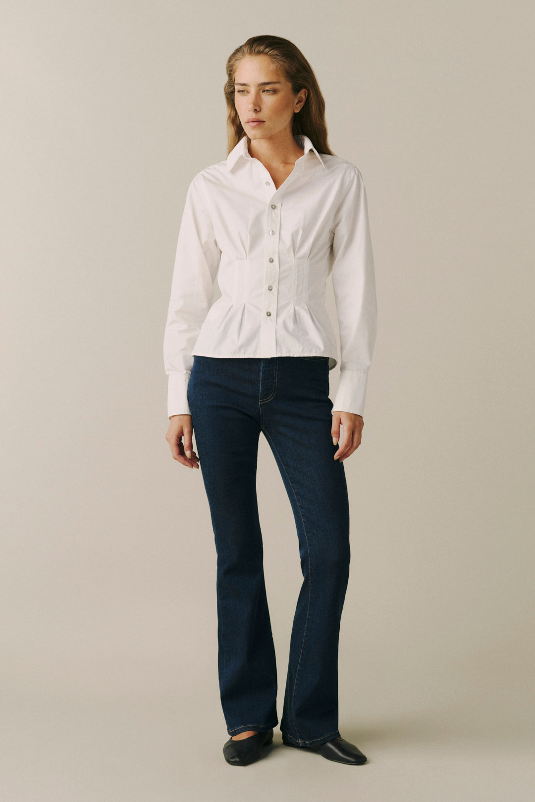 A woman modeling a white long-sleeve button-up shirt with structured shoulders and dark blue flared jeans against a plain beige studio background.
