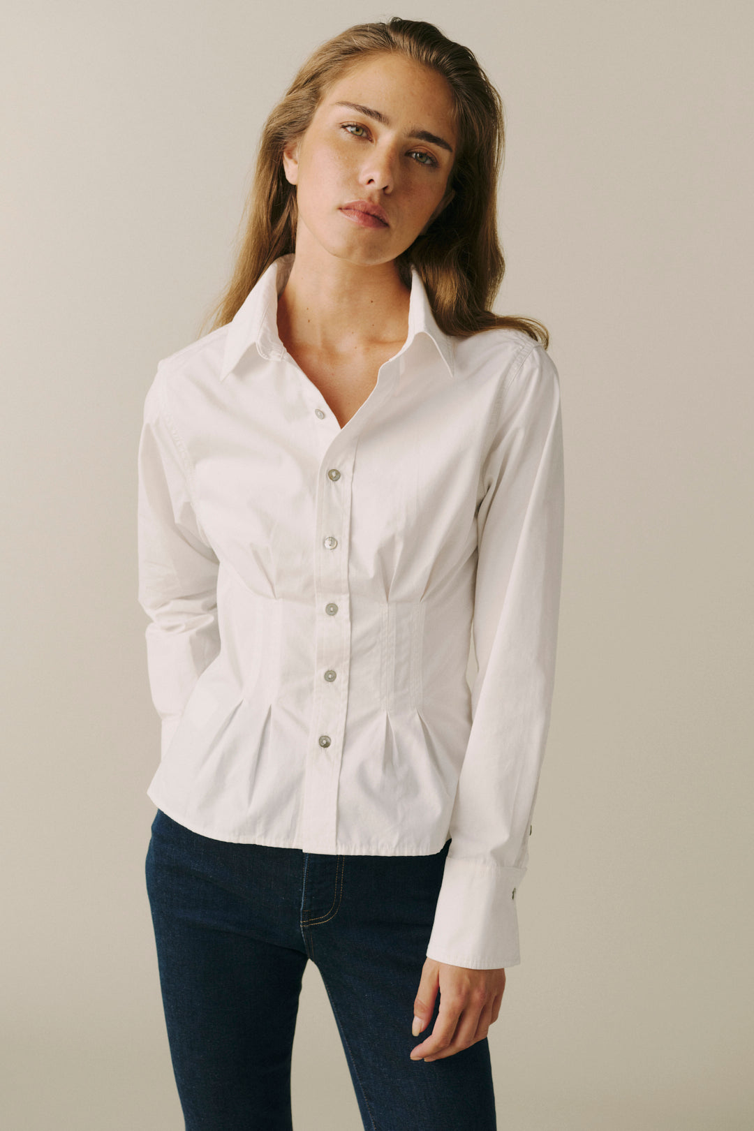 A woman modeling a white long-sleeve button-up shirt with a structured, slightly gathered waist and dark blue jeans, set against a plain beige studio background.