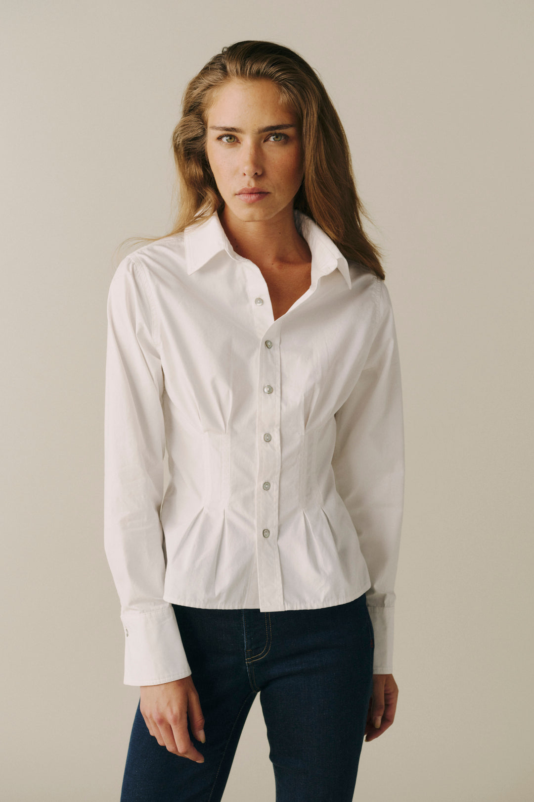 A woman modeling a white long-sleeve button-up shirt with a structured collar and a subtle peplum waist, paired with dark blue jeans, against a solid light beige background.
