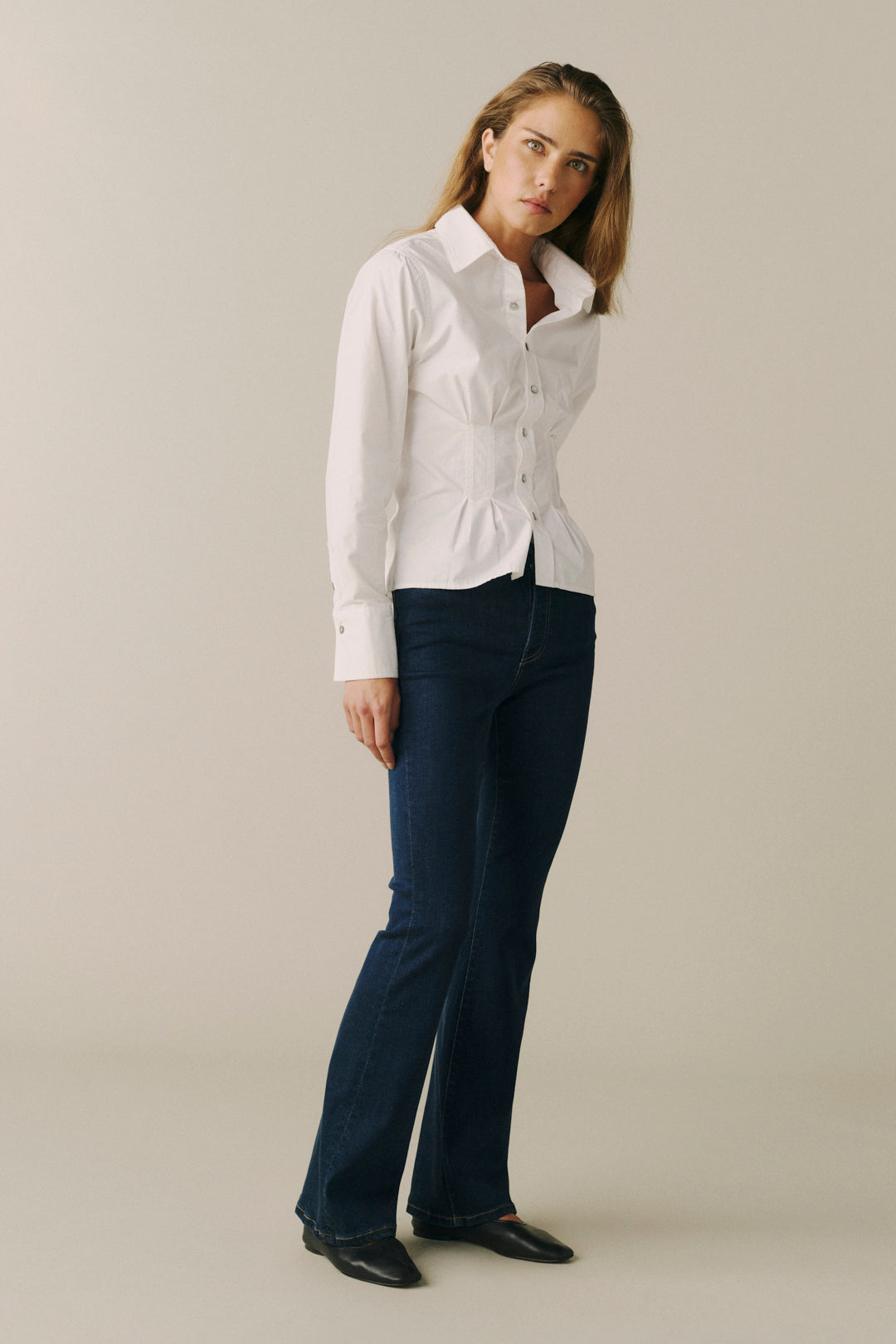 A woman modeling a white long-sleeve button-up shirt tucked into dark blue flared jeans, standing against a plain light beige studio background.
