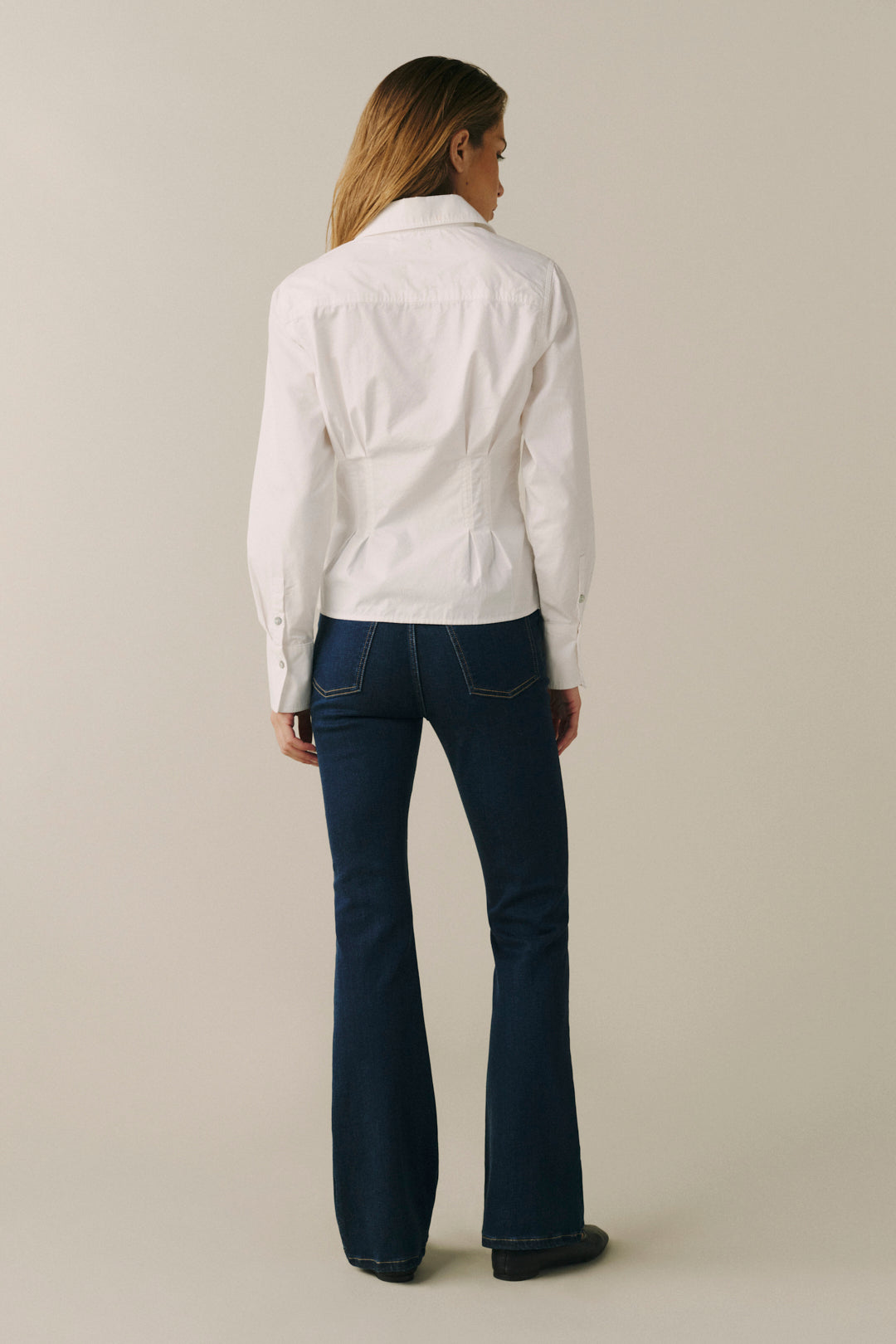 A woman shown from the back wearing a white long-sleeve button-up shirt and dark blue flared jeans against a plain light beige studio background.
