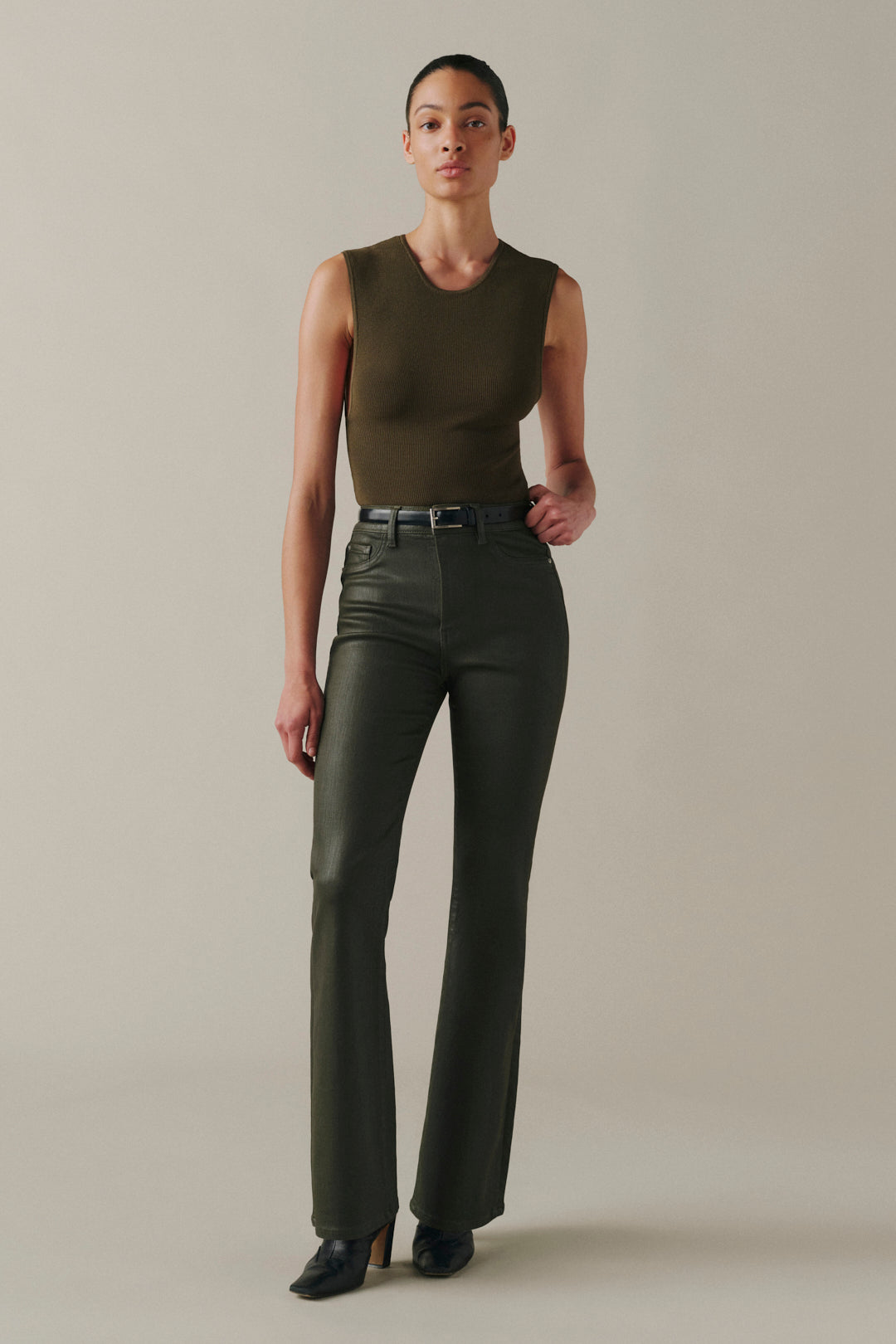 A model stands against a plain beige studio background wearing a sleeveless olive-brown top and dark olive green high-waisted flared pants with a black belt.