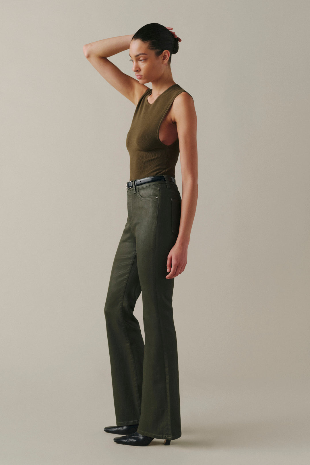 A model is shown in profile wearing a sleeveless olive green top and matching olive green wide-leg pants, paired with a black belt and black shoes, against a plain beige studio background.