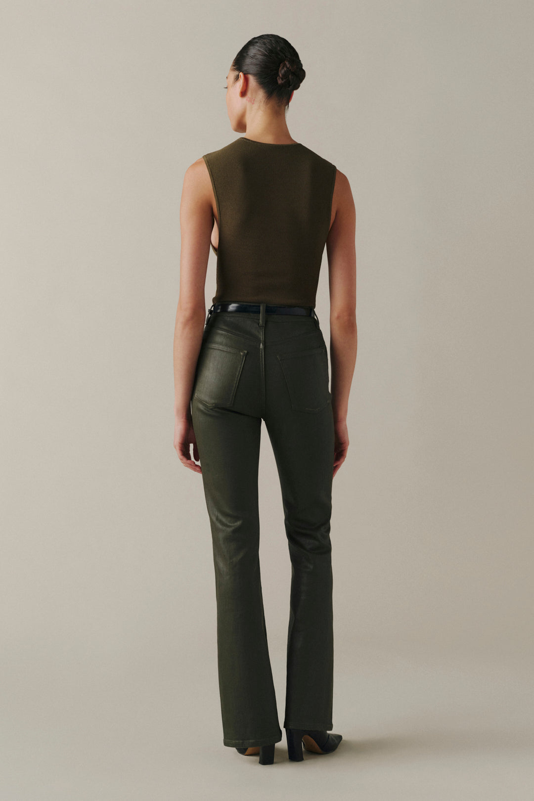 A model shown from the back wearing dark olive green leather pants and a matching sleeveless top, set against a plain light gray studio background.