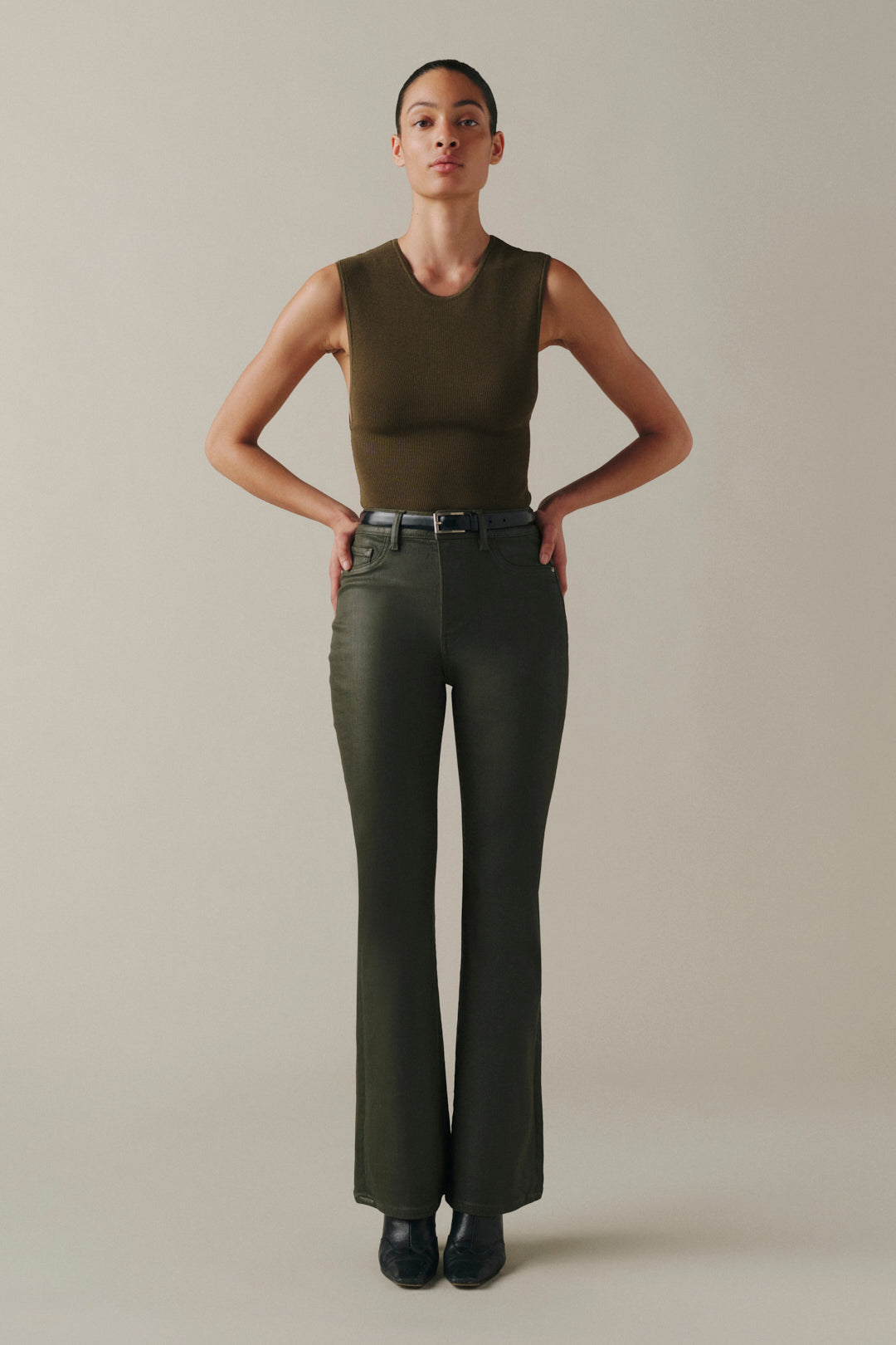 A model stands against a plain beige studio background wearing an olive green sleeveless top and matching olive green pants with a dark belt and black shoes.