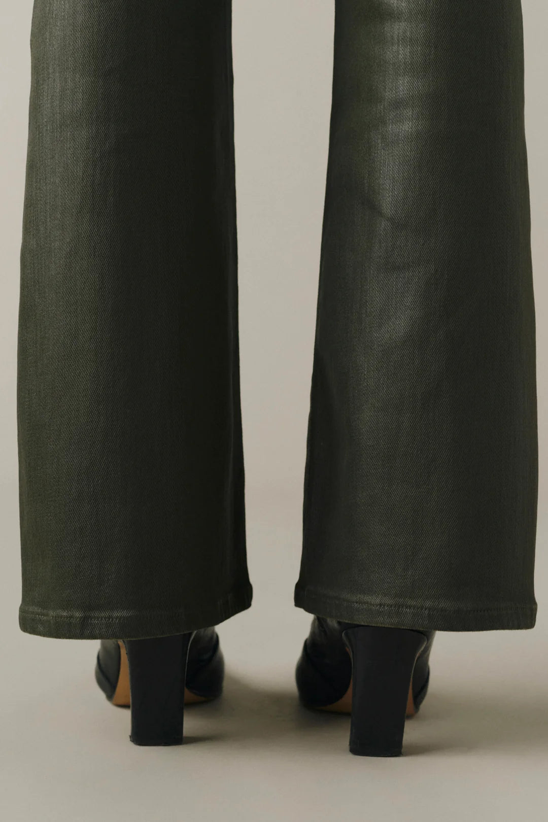 Close-up rear view of a person wearing dark olive green wide-leg jeans and black block-heel shoes against a plain light gray studio background.