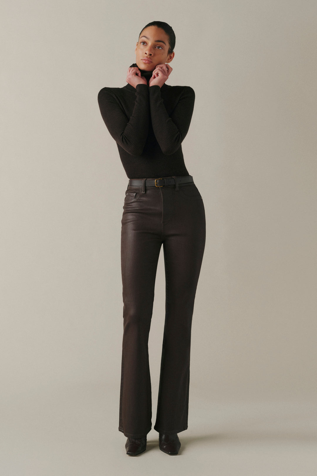 A model wearing a black long-sleeve top and dark brown high-waisted leather-look flare pants with a black belt, standing against a neutral beige studio background.