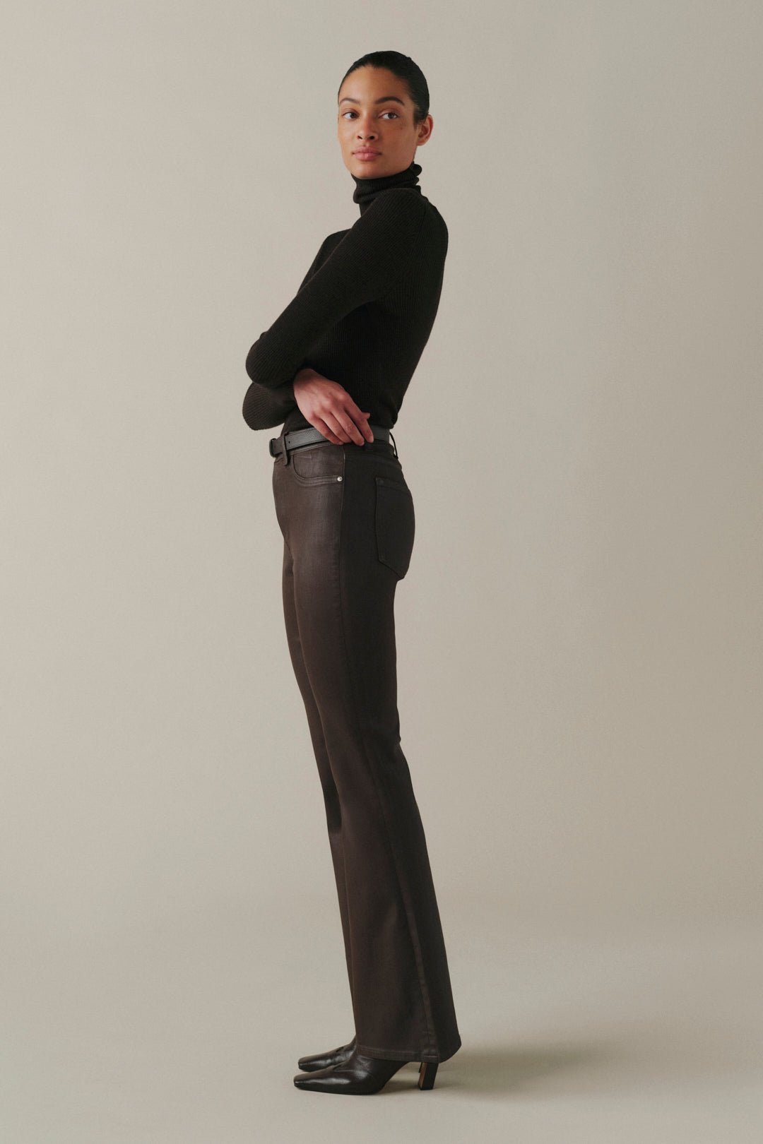 A model is shown in a side profile wearing a black turtleneck sweater and dark brown leather trousers, standing against a solid light beige studio background.