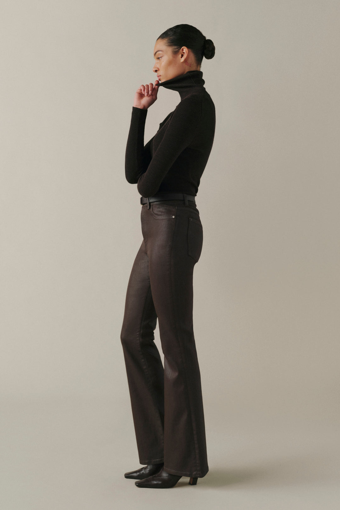 A model in side profile wearing a black long-sleeve turtleneck top and dark brown leather flare pants, standing against a neutral light gray studio background.
