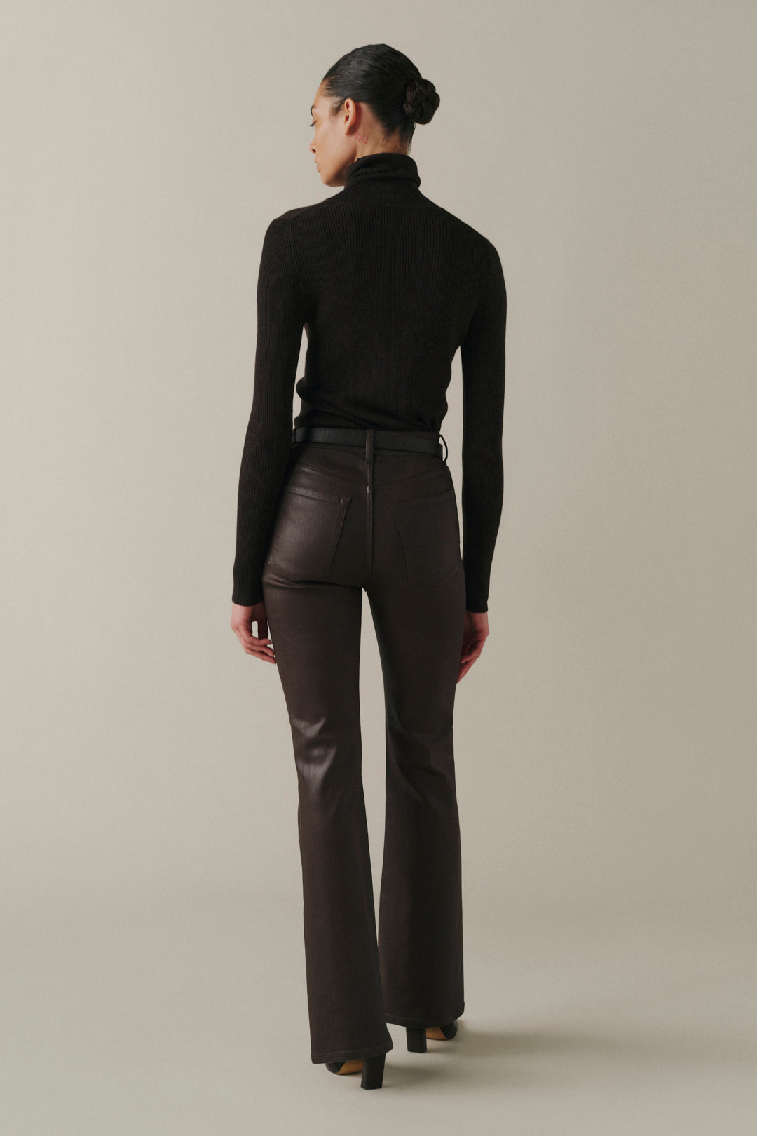 A woman is shown from behind wearing a black long-sleeve turtleneck sweater and dark brown leather flare pants against a neutral beige studio background.