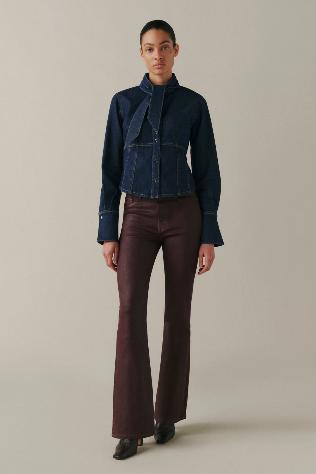 A model stands against a plain beige studio background wearing a dark blue denim jacket with a high structured collar and a draped tie detail at the chest, paired with dark brown flared trousers and black shoes.