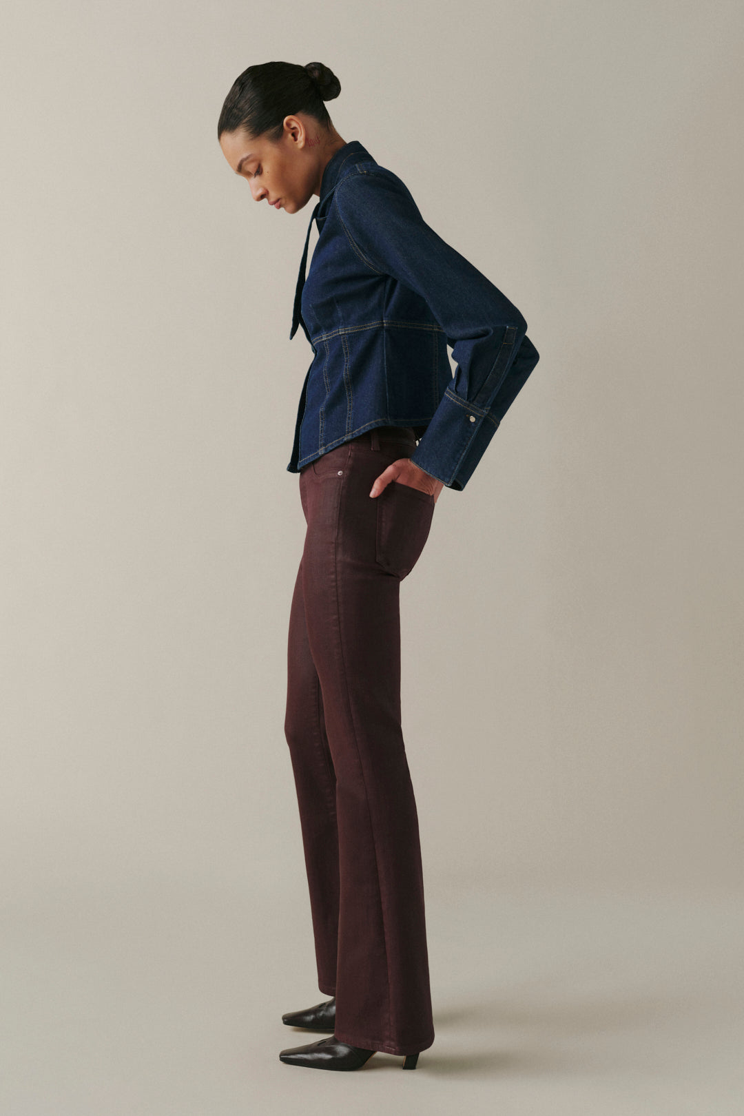 A model is shown in profile wearing a dark blue cropped denim jacket and dark brown wide-leg trousers, standing against a solid light beige studio background.