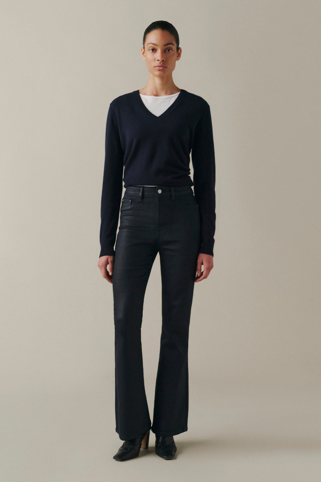 A model stands against a plain beige studio background wearing a dark navy long-sleeve V-neck sweater over a white crew-neck top, paired with dark charcoal flared jeans and black pointed-toe heels.
