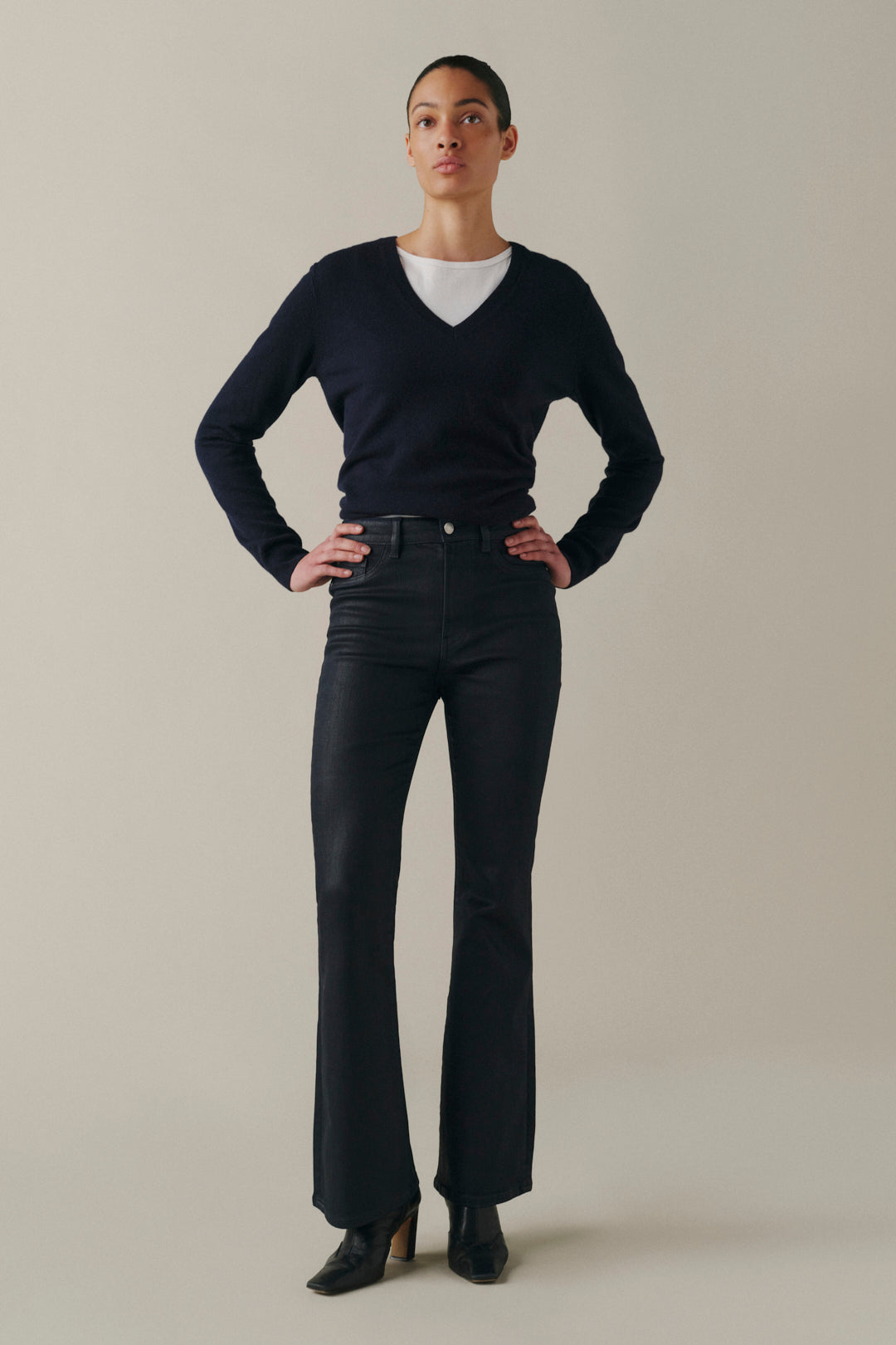 A model stands against a plain beige studio background wearing a dark navy long-sleeve V-neck sweater over a white crew-neck top, paired with dark charcoal flared jeans and black pointed-toe heels.