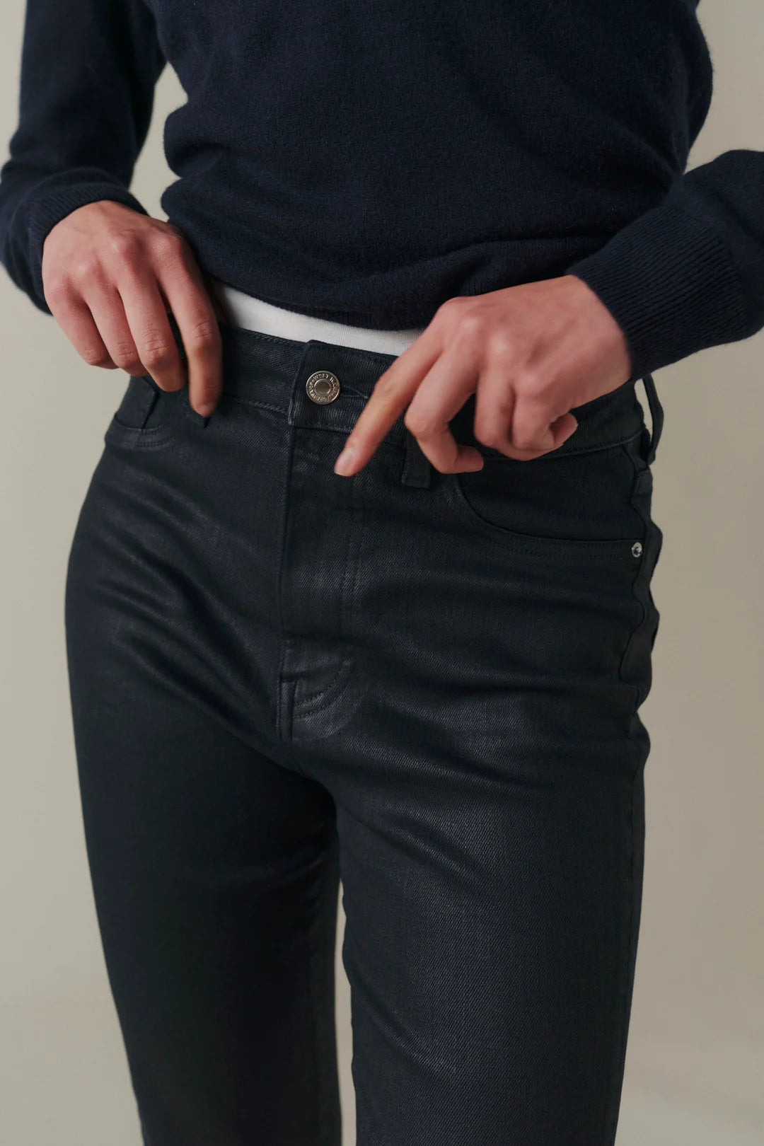 Close-up of a person wearing dark charcoal gray slim-fit jeans, pulling the waistband slightly to show the fit and button area, with a dark navy sweater visible above.