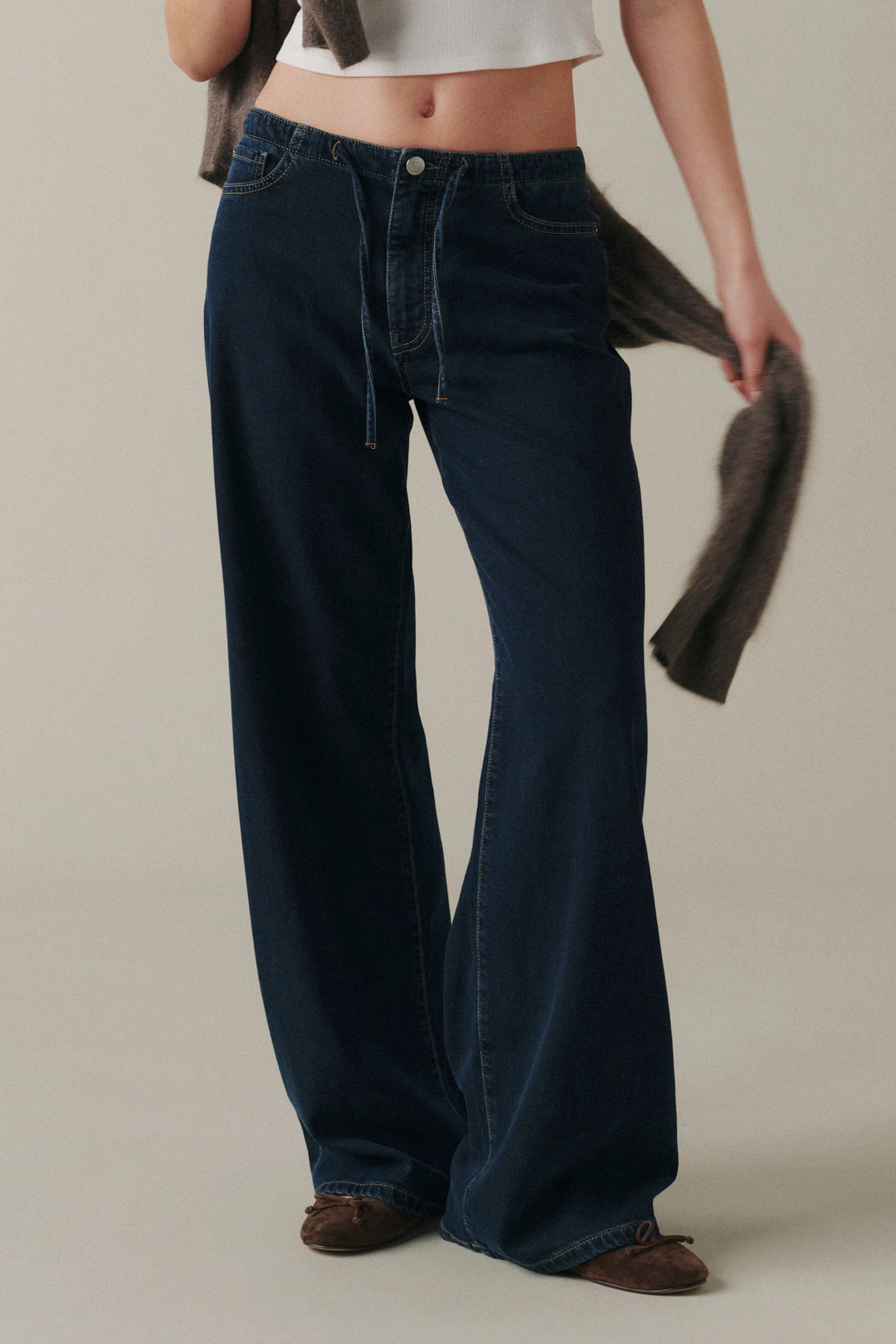 A model wearing dark blue wide-leg denim jeans, styled with a white crop top and brown shoes, holding a brown jacket against a neutral beige background.