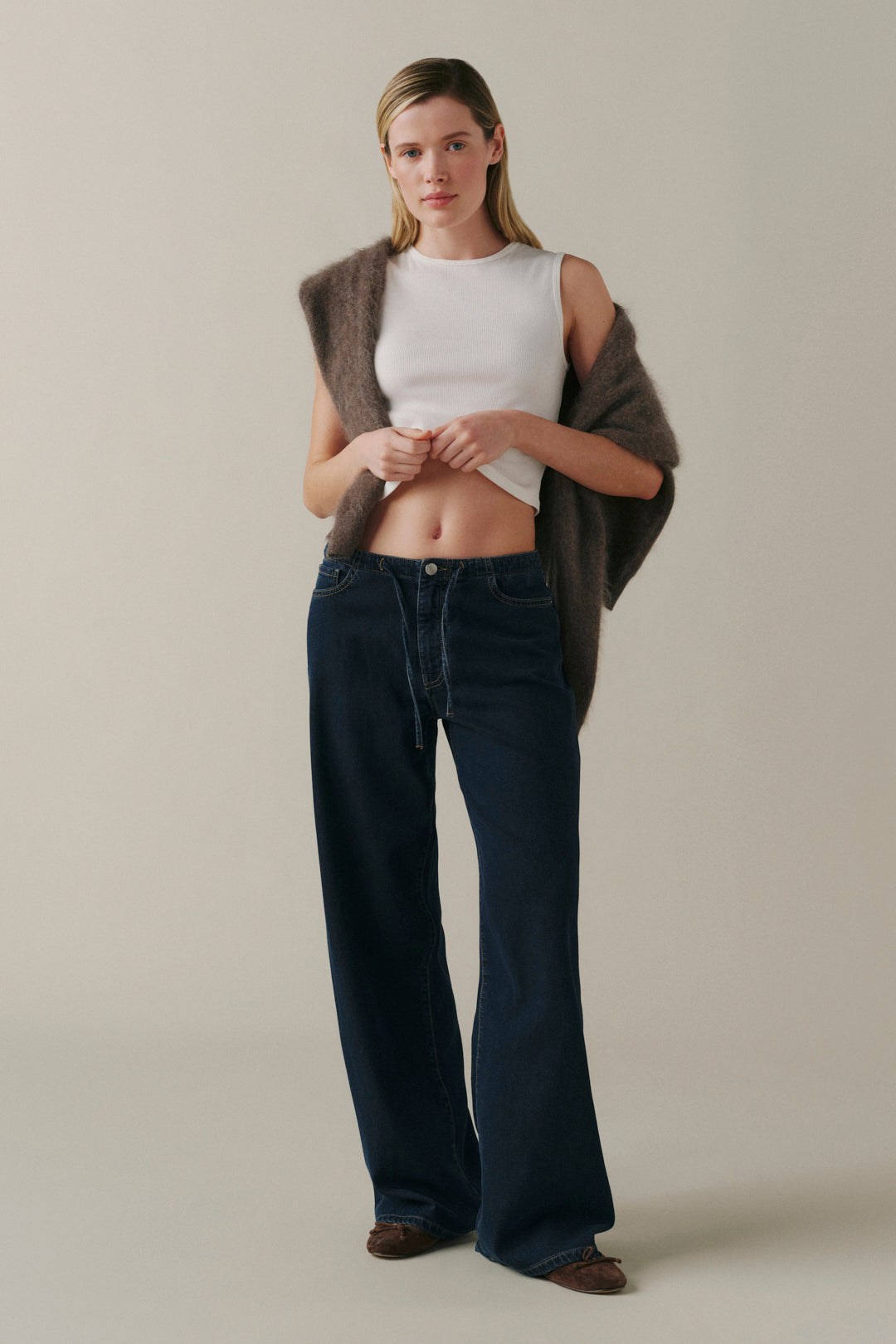 A model stands against a plain beige studio background wearing dark blue wide-leg jeans, a white cropped top, and a brown fuzzy sleeveless cardigan.