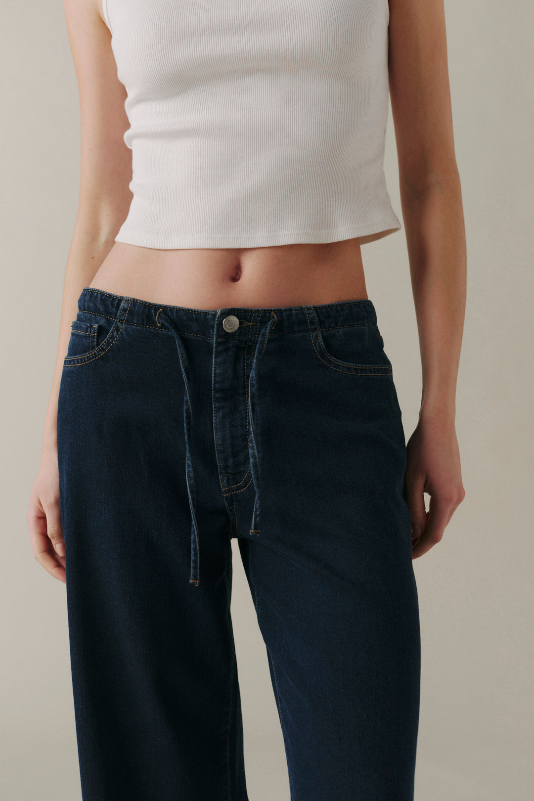 A person wearing dark blue wide-leg jeans with a drawstring waistband and a white cropped tank top, shown from the chest down against a plain beige studio background.