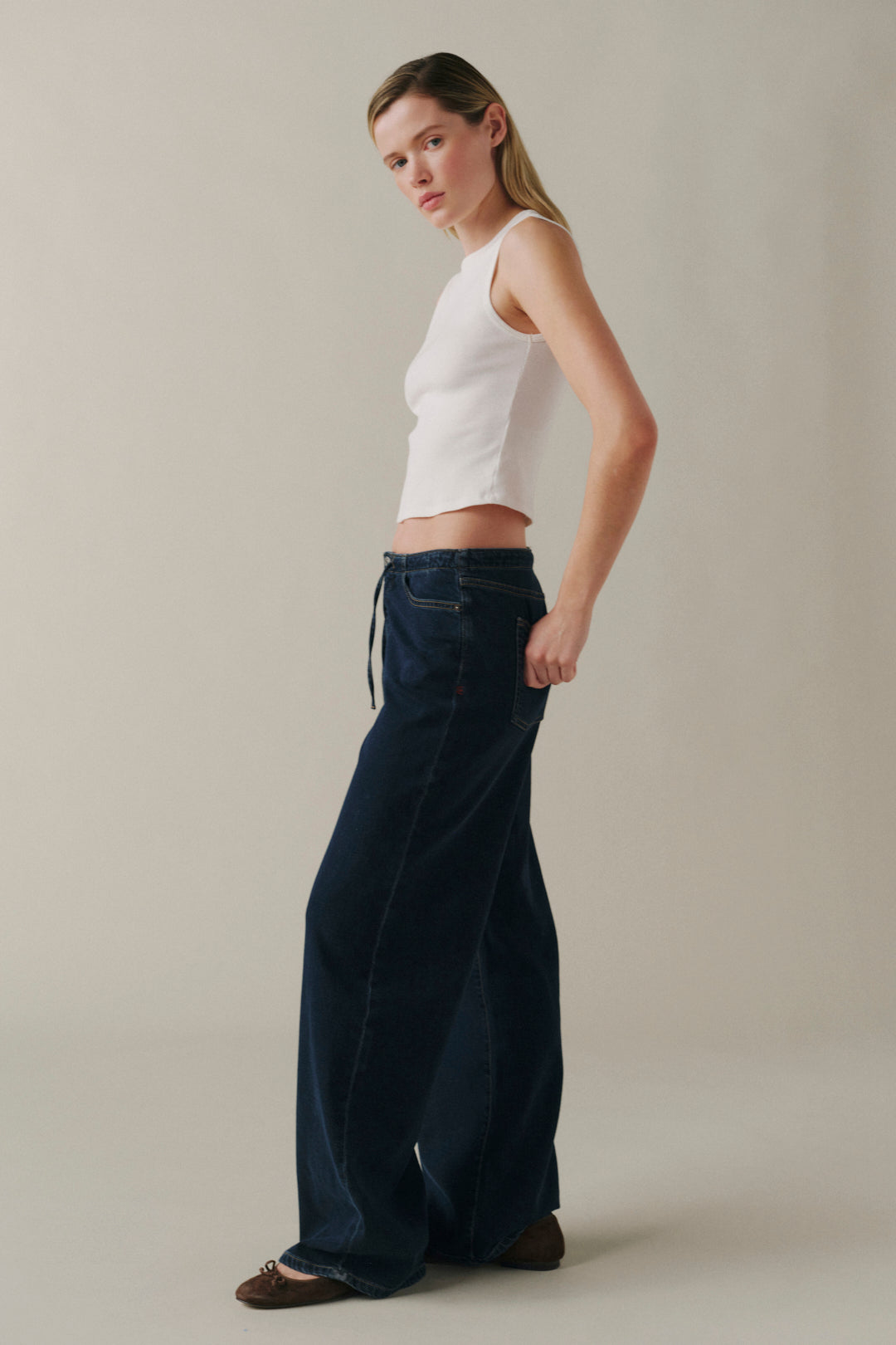 A woman modeling dark blue wide-leg jeans with a drawstring waist, paired with a white ribbed crop top, standing against a neutral beige studio background.