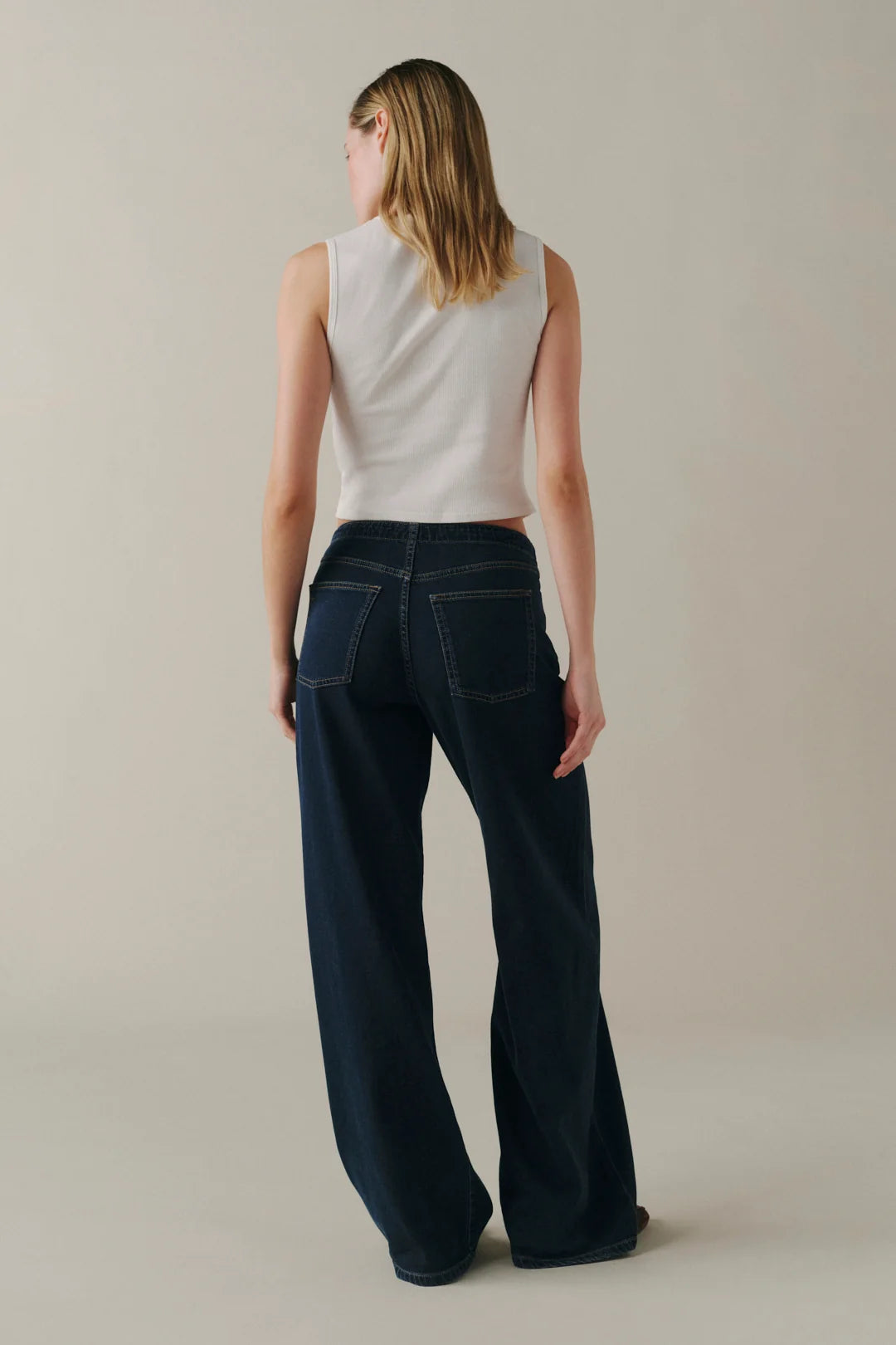 A woman shown from the back wearing dark wide-leg jeans and a white sleeveless top, standing against a plain light beige studio background.