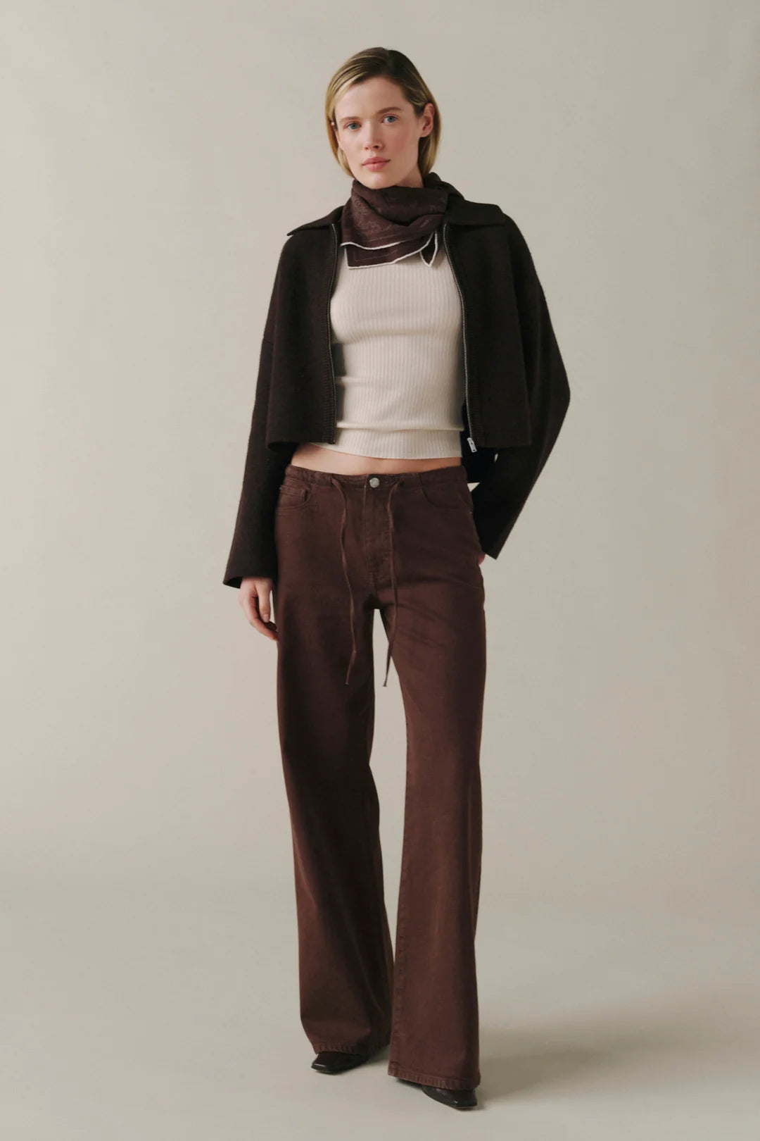 A model stands against a plain light beige studio background wearing a dark brown cropped jacket, cream ribbed top, dark brown scarf, and dark brown wide-leg pants.