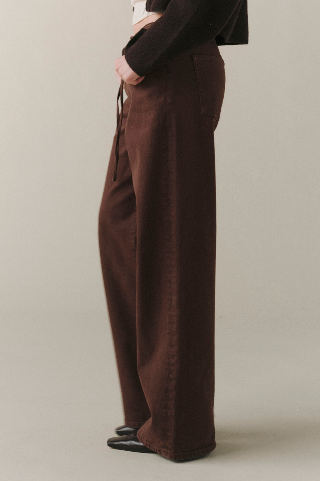 A person shown from the waist down wearing dark brown wide-leg pants, a black long-sleeve top, and black shoes, standing against a plain light beige studio background.
