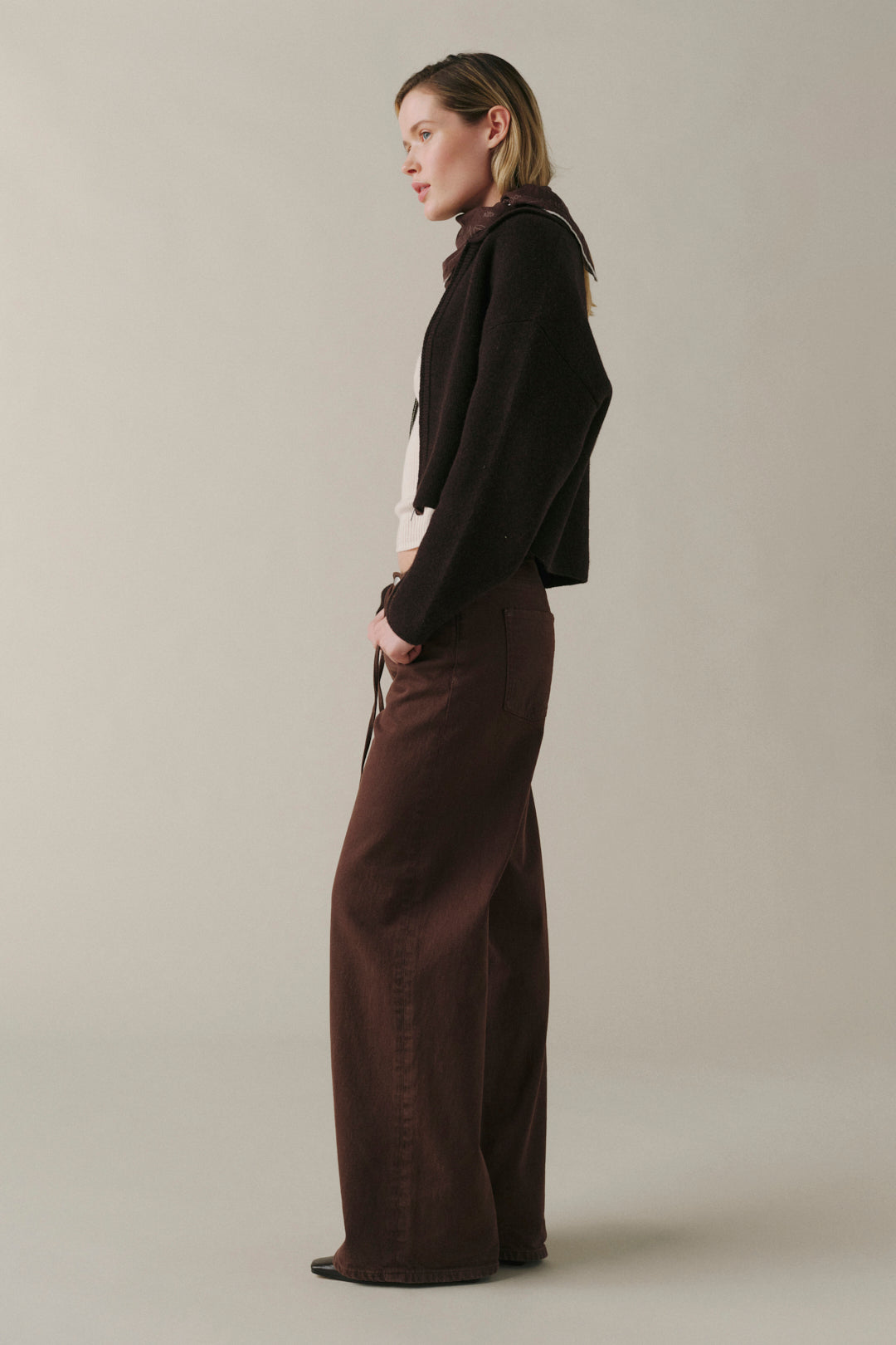A woman stands in profile wearing a dark cropped jacket, a white top, and wide-leg brown trousers against a plain light gray studio background.