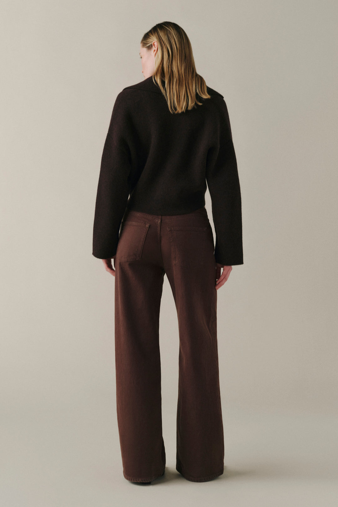 A model shown from the back wearing a dark brown long-sleeve knit top and matching dark brown wide-leg trousers, set against a plain light gray studio background.