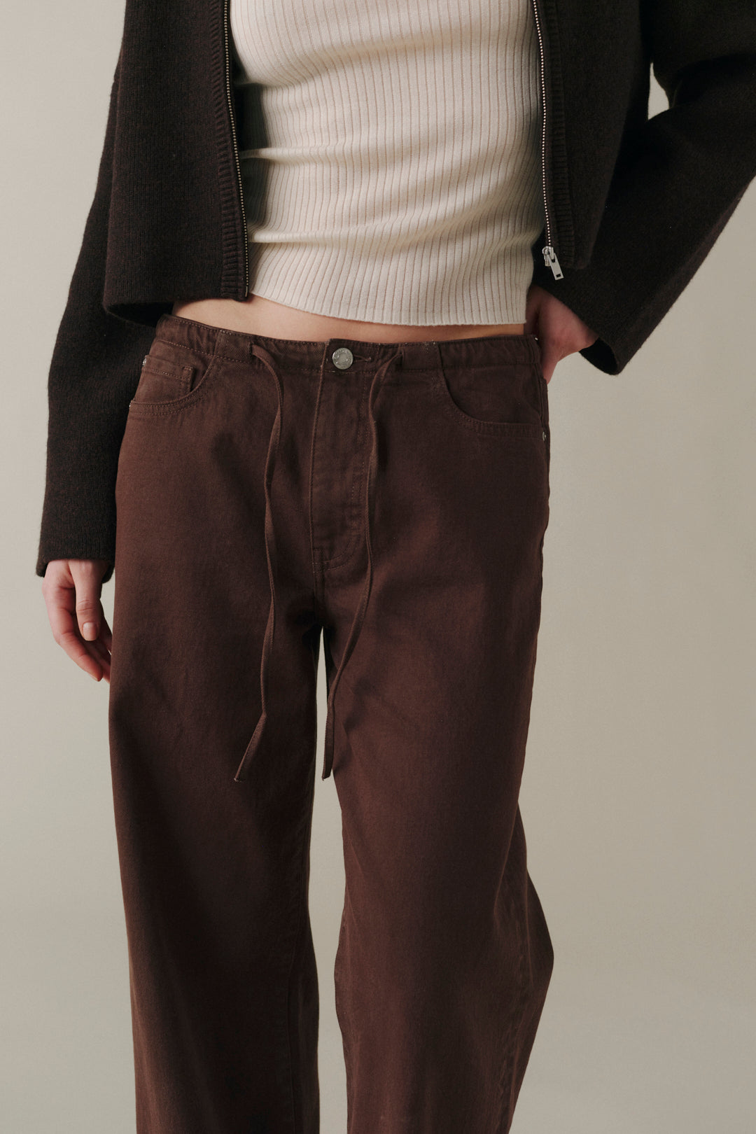 A person wearing dark brown wide-leg drawstring pants, styled with a cream ribbed knit top and a dark brown cropped jacket, against a neutral beige background.