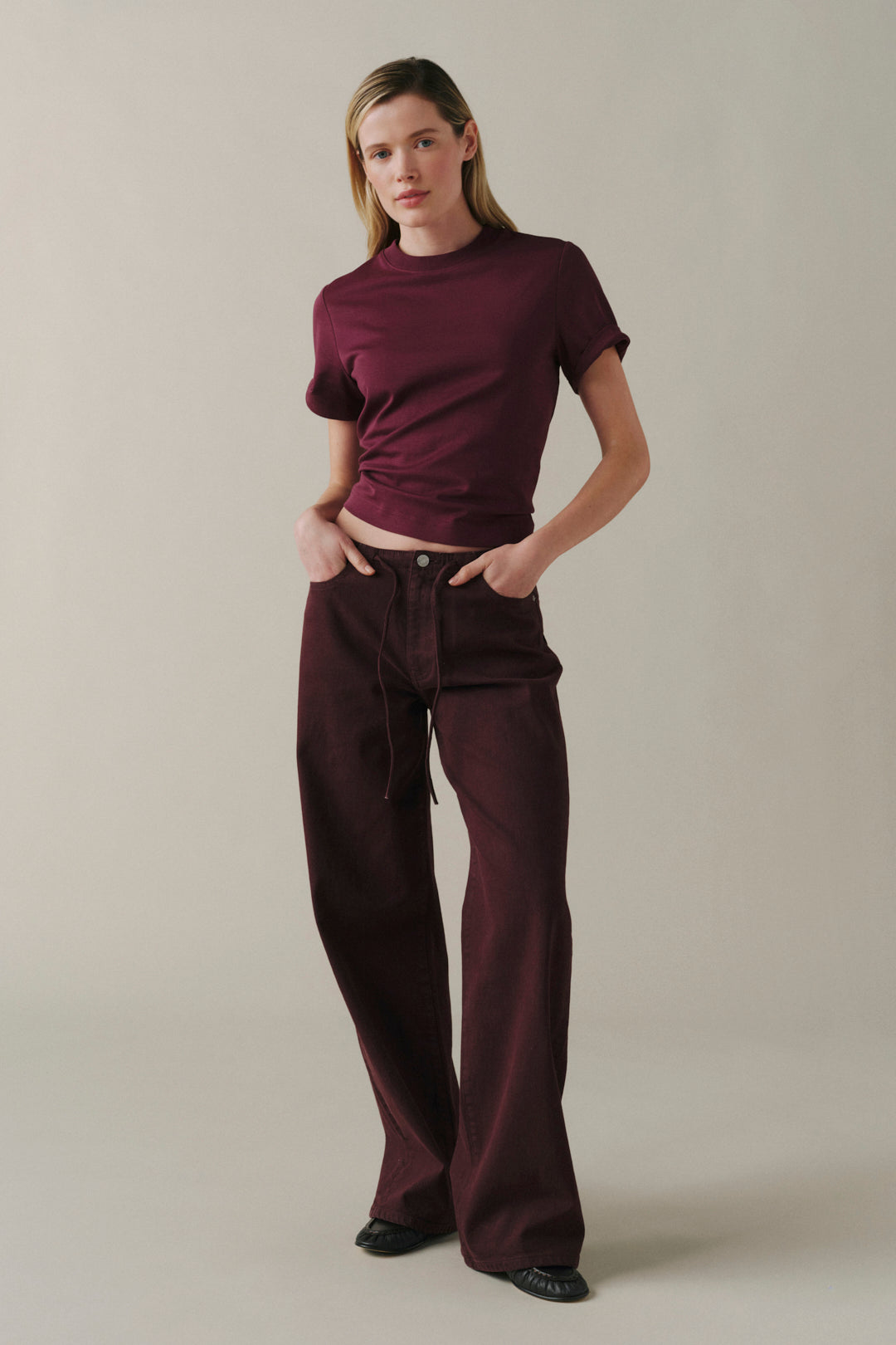 A woman modeling a matching burgundy short-sleeve crop top and wide-leg drawstring pants against a plain beige studio background.