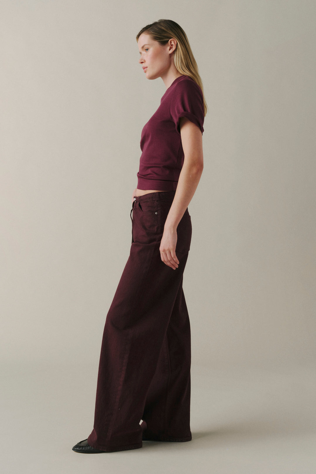 A woman in profile wearing a short-sleeve maroon crop top and matching maroon wide-leg pants, standing against a plain light beige studio background.