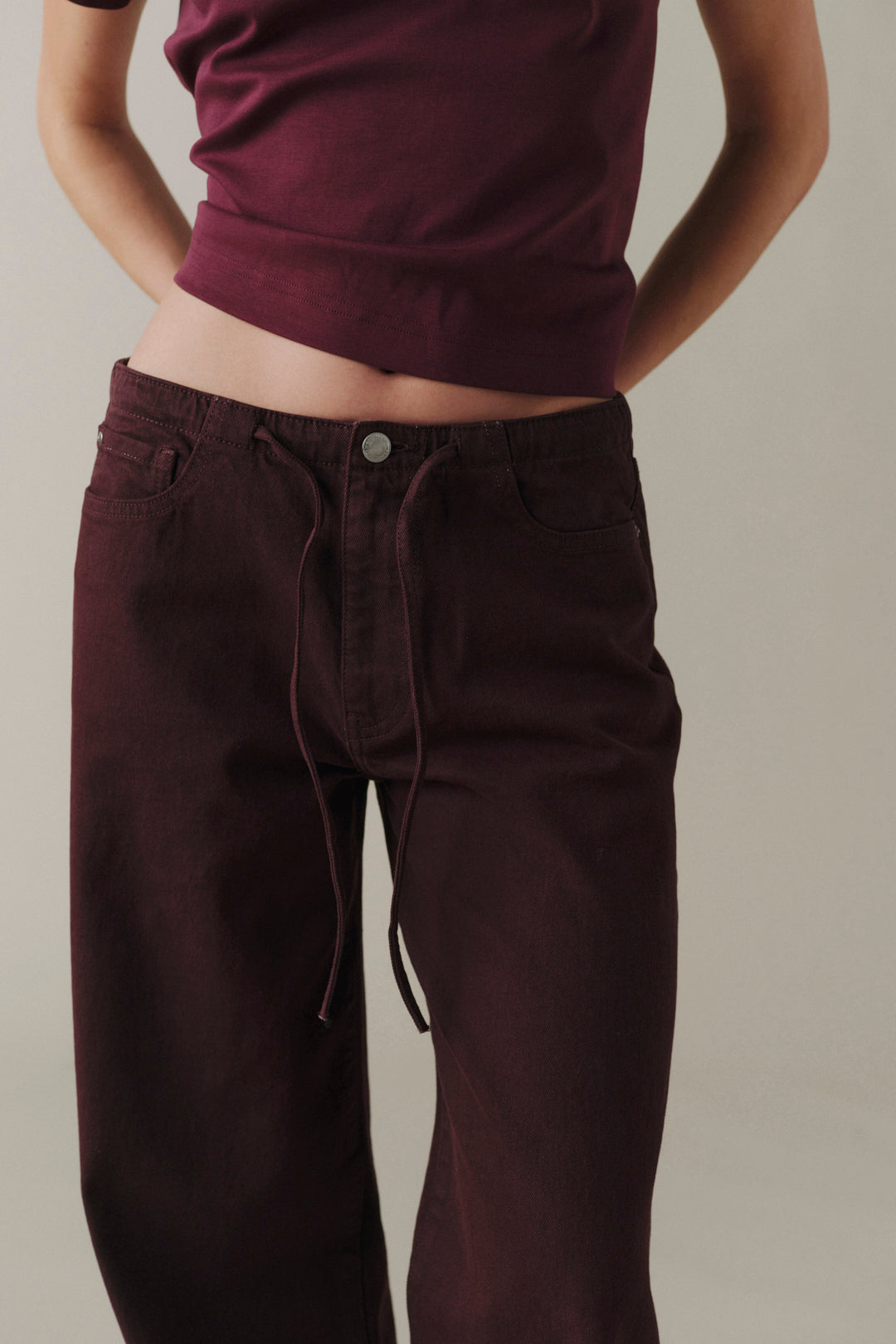 A person wearing dark burgundy drawstring pants and a matching dark burgundy cropped top, shown from the torso down against a plain light beige studio background.