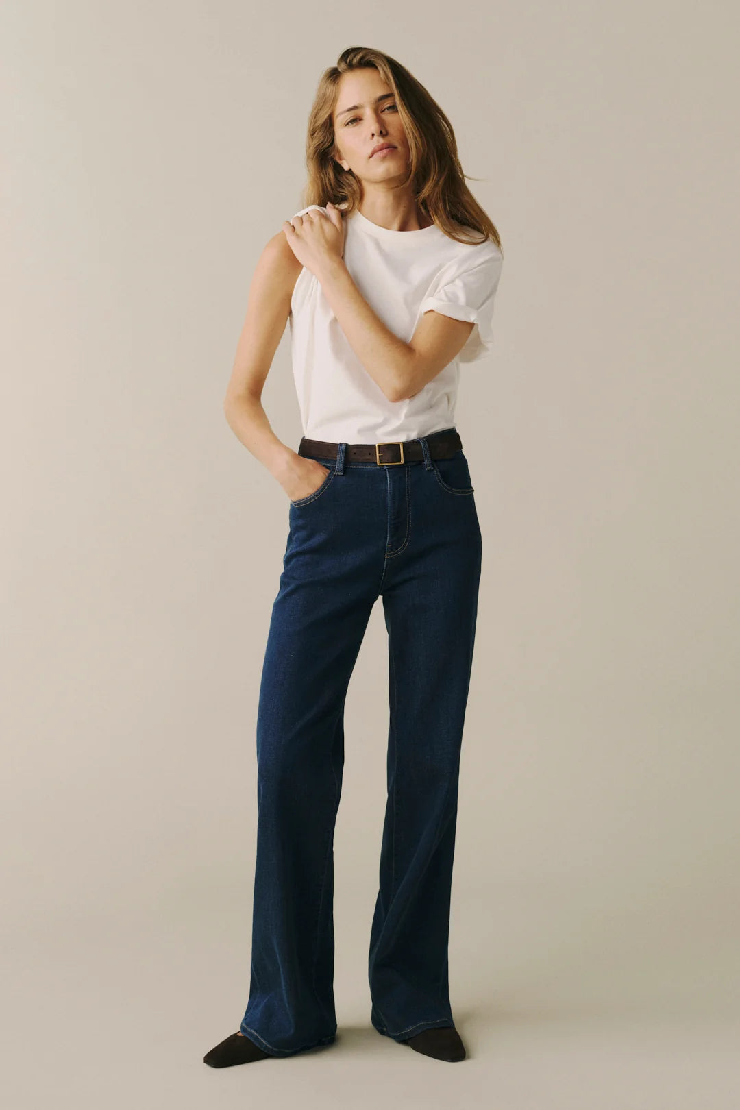 A woman modeling a pair of dark blue wide-leg jeans with a white t-shirt and a brown belt, standing against a plain beige studio background.