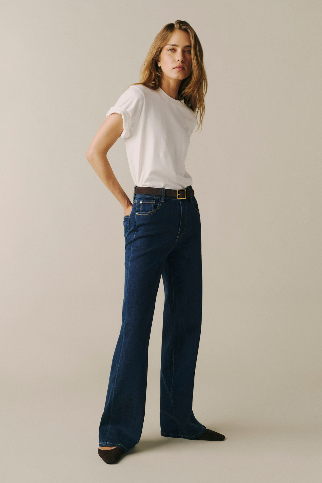 A woman modeling dark blue wide-leg jeans with a brown belt, paired with a white t-shirt, standing against a solid light beige background.