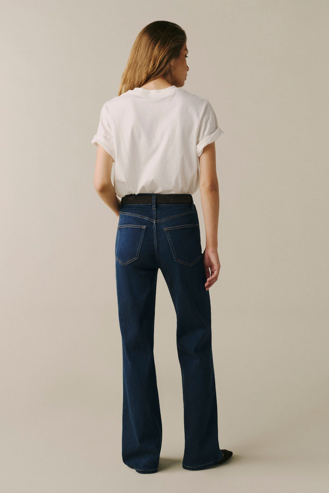A woman viewed from behind wearing dark blue wide-leg jeans, a black belt, and a white t-shirt, standing against a solid light beige background.