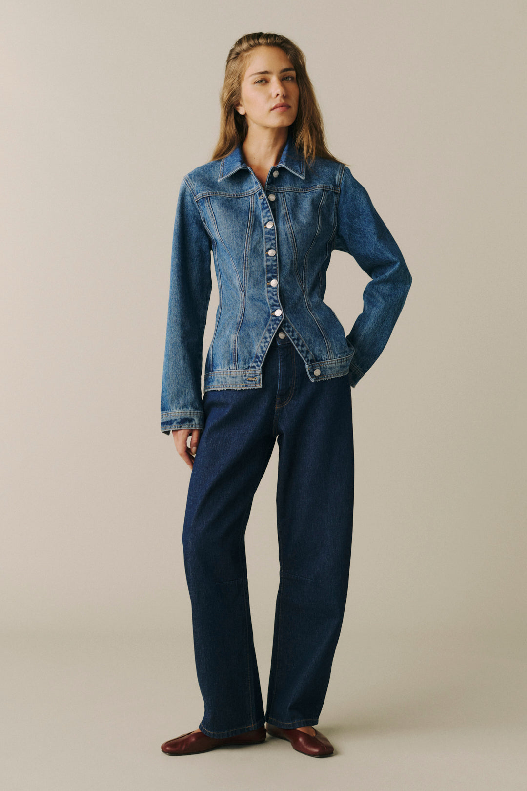 A woman modeling a blue denim jacket with front buttons and a cropped hem, paired with dark blue wide-leg jeans and brown shoes, against a plain beige studio background.
