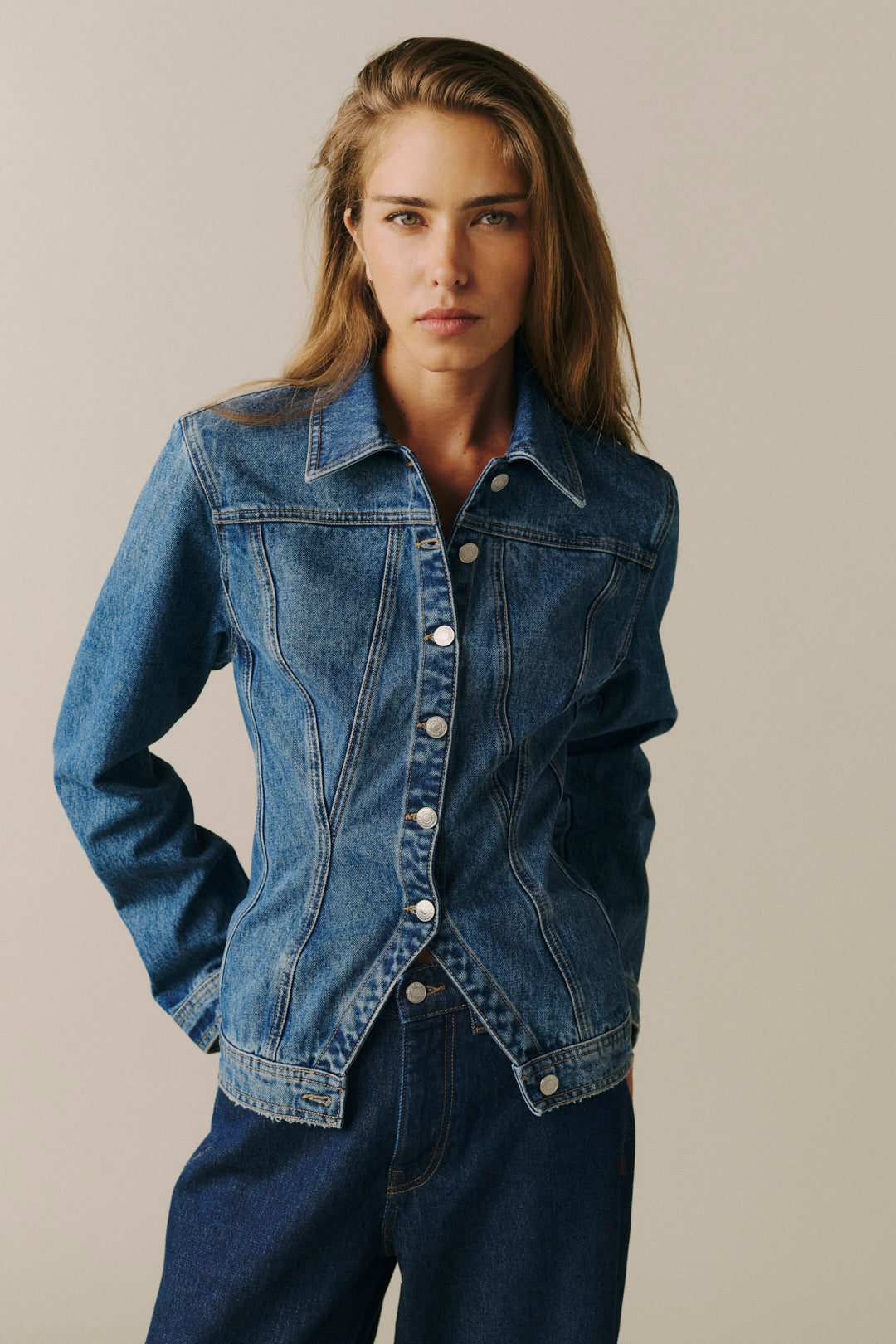 A woman wearing a blue denim jacket with front button closure and matching blue jeans, standing against a plain light beige studio background.