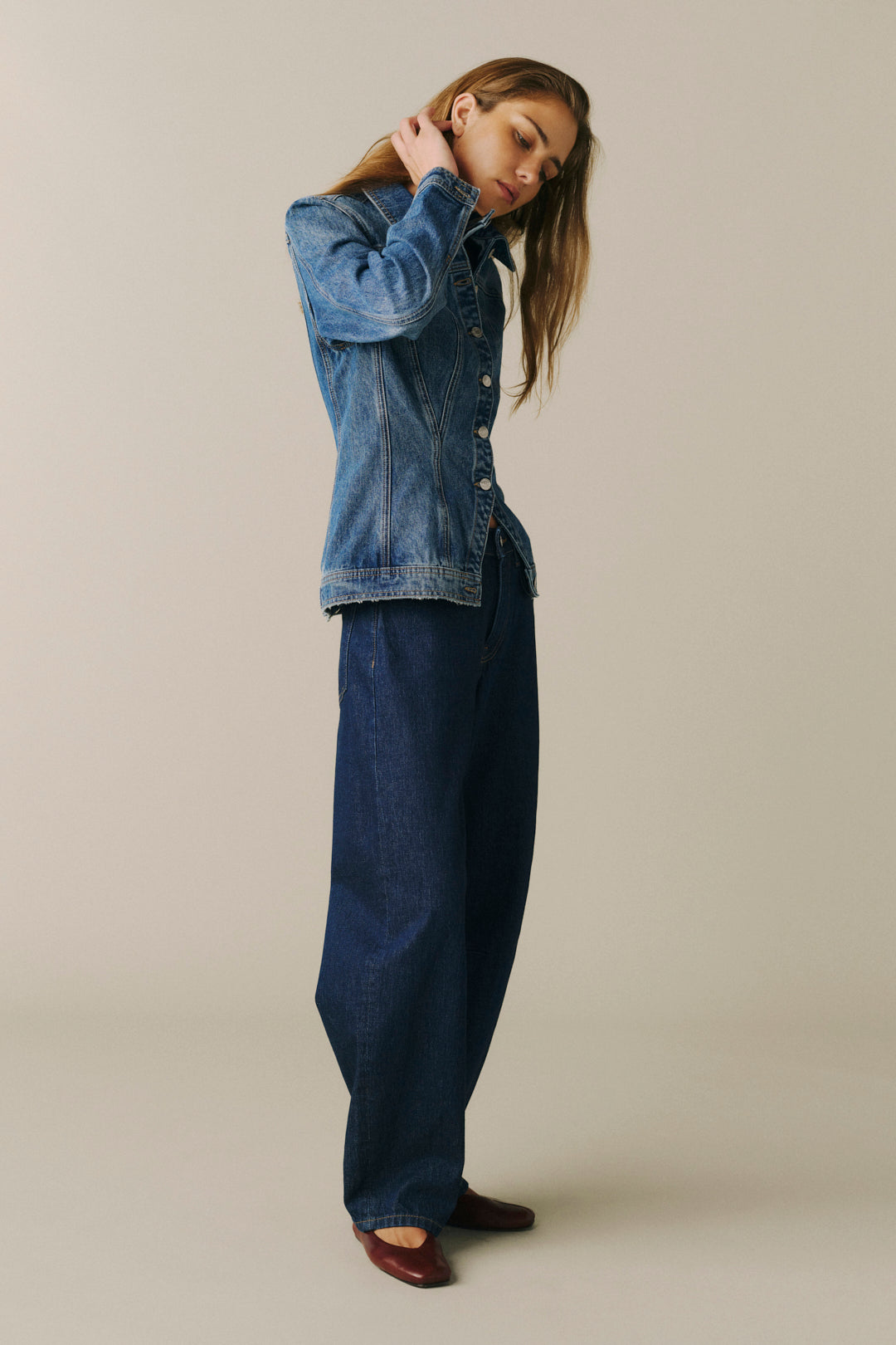 A woman modeling a blue denim jacket and matching dark blue denim pants against a plain beige studio background.