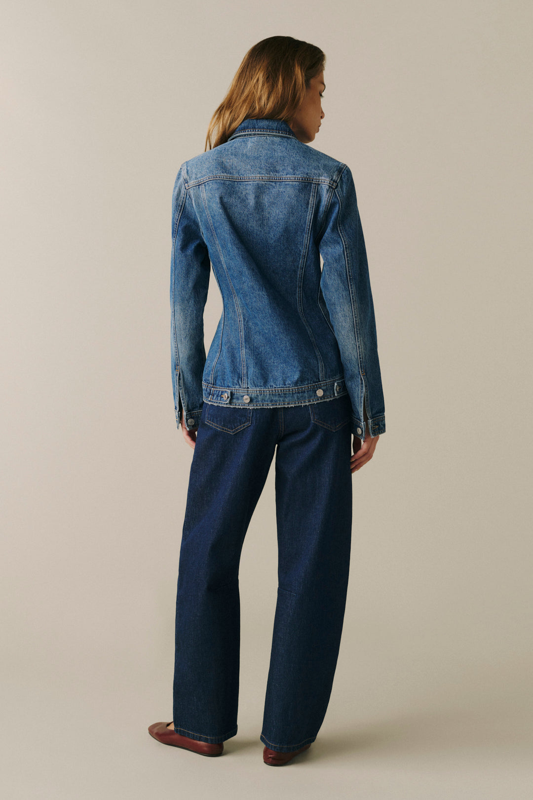 A woman shown from the back wearing a medium-wash blue denim jacket and dark blue jeans, standing against a plain light beige studio background.