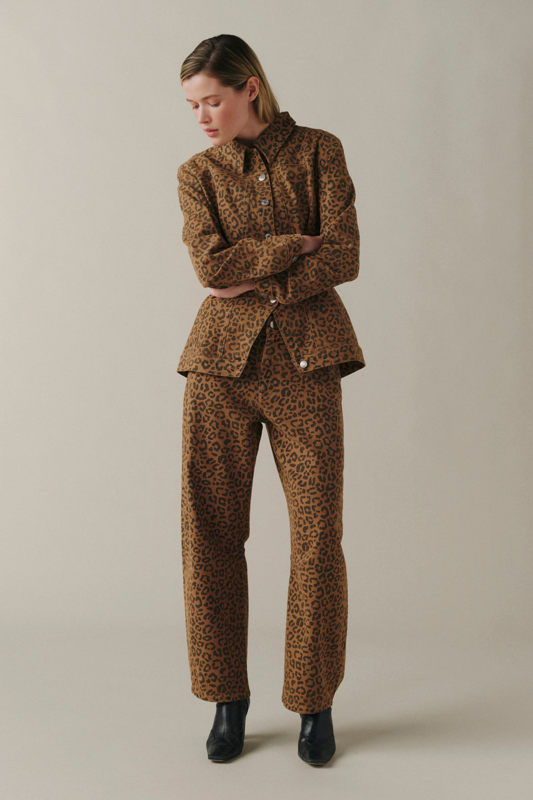 A model wearing a matching brown and black leopard-print jacket and trousers set, styled with black boots, against a solid light beige studio background.