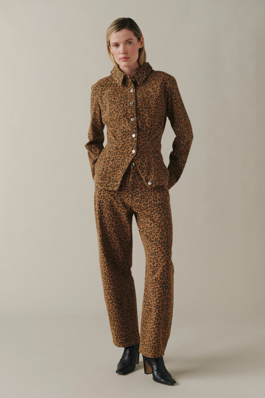 A model wearing a matching brown and black leopard-print blazer and trousers suit, paired with black ankle boots, standing against a neutral beige studio background.