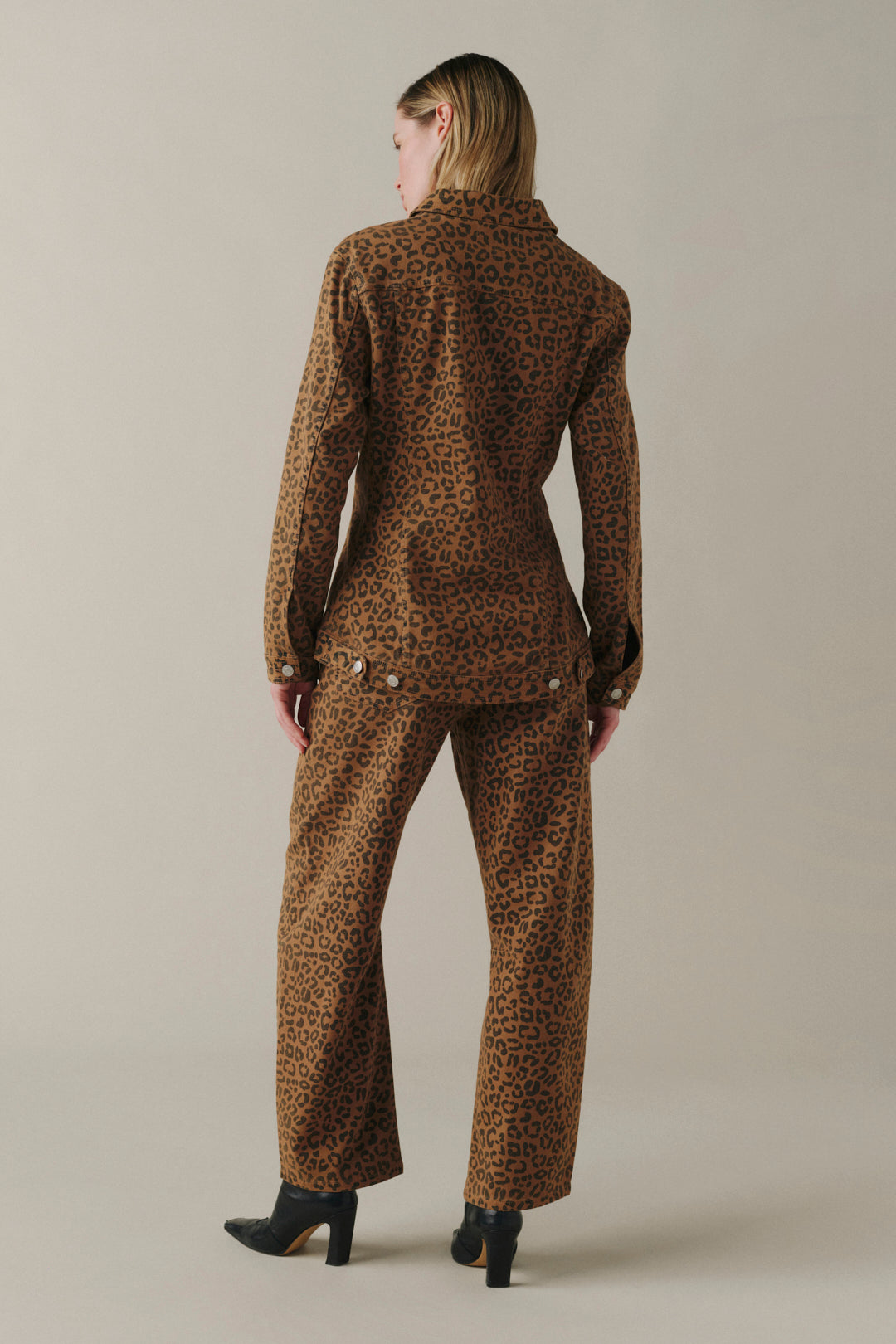 A model shown from the back wearing a matching brown and black leopard-print blazer and trousers set, paired with black high-heeled boots, against a solid light beige studio background.