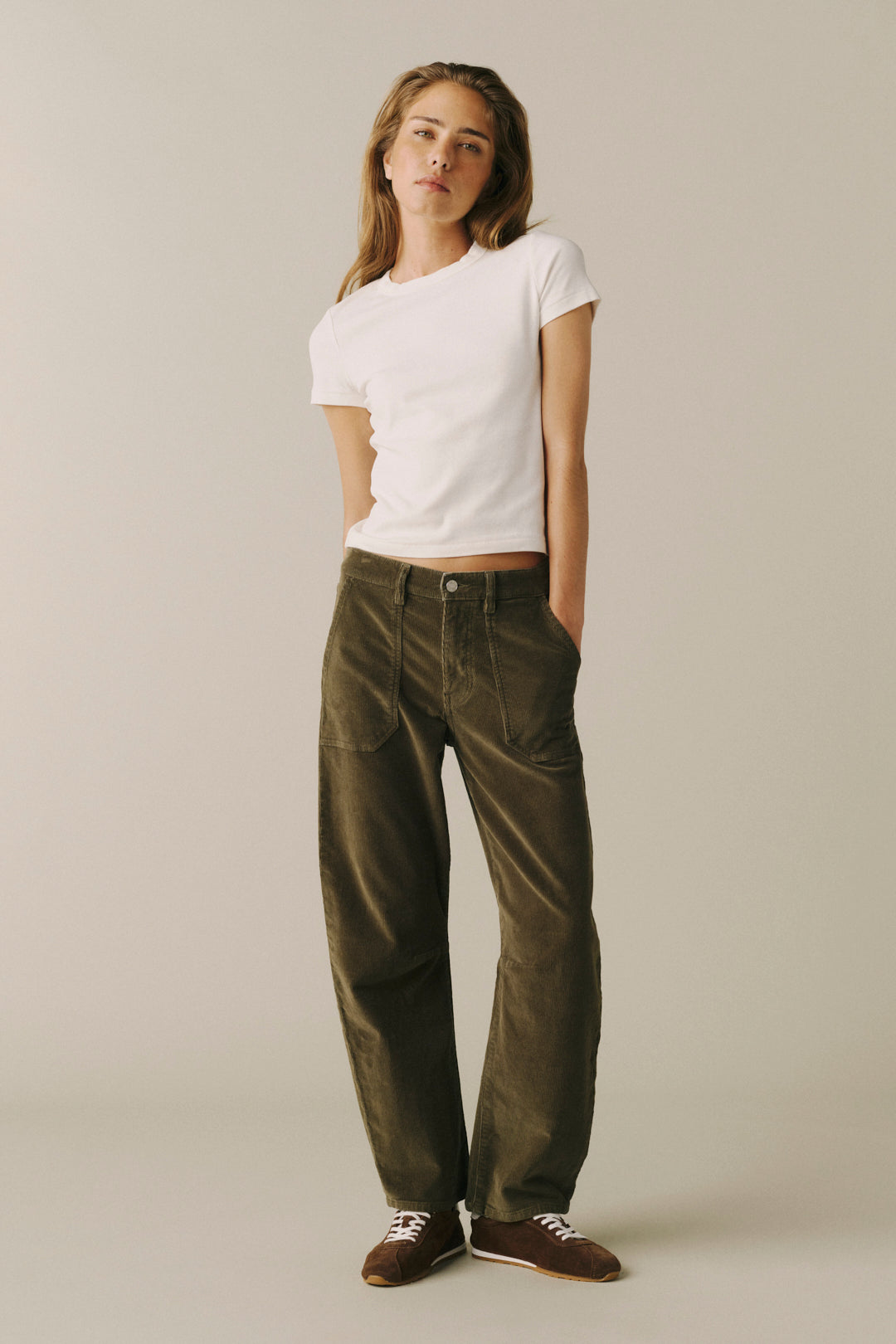 A woman modeling a white short-sleeve t-shirt and dark olive green wide-leg corduroy pants, paired with brown leather sneakers, against a plain light gray studio background.